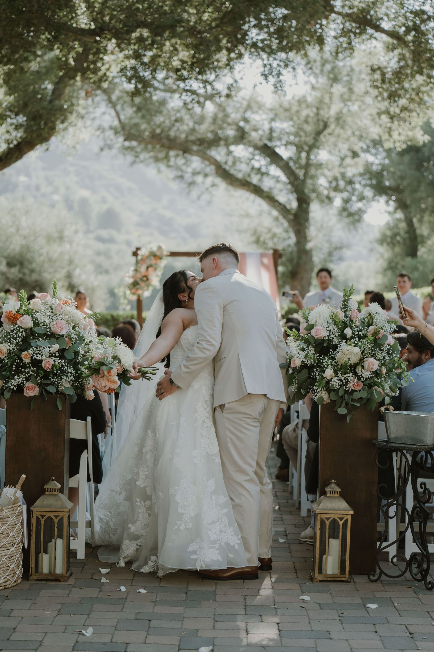 Bride and groom share first kiss at outdoor wedding ceremony aisle lined with floral arrangements and lanterns.