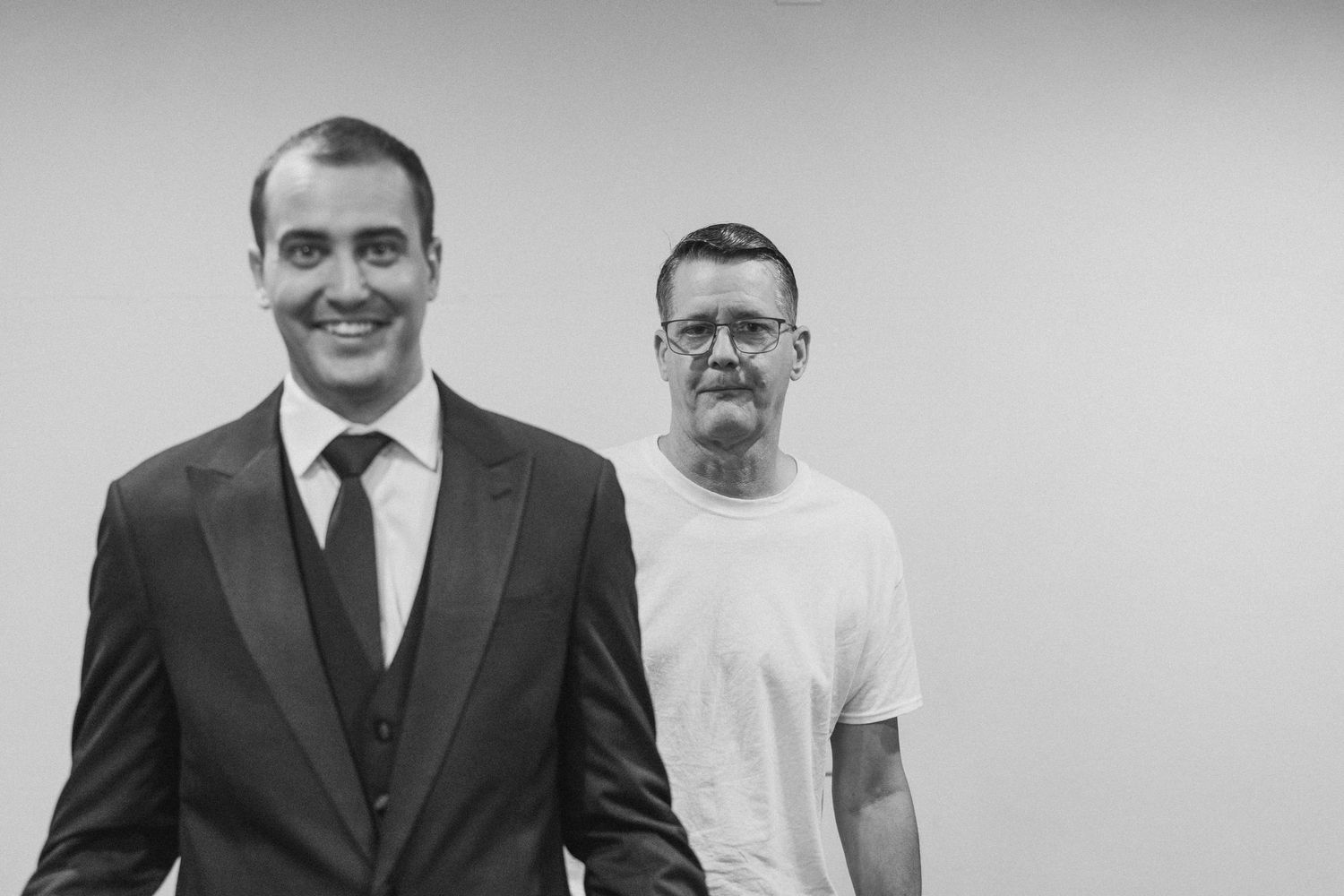 Two men posing in black and white photo, one in suit and tie, other in white t-shirt.