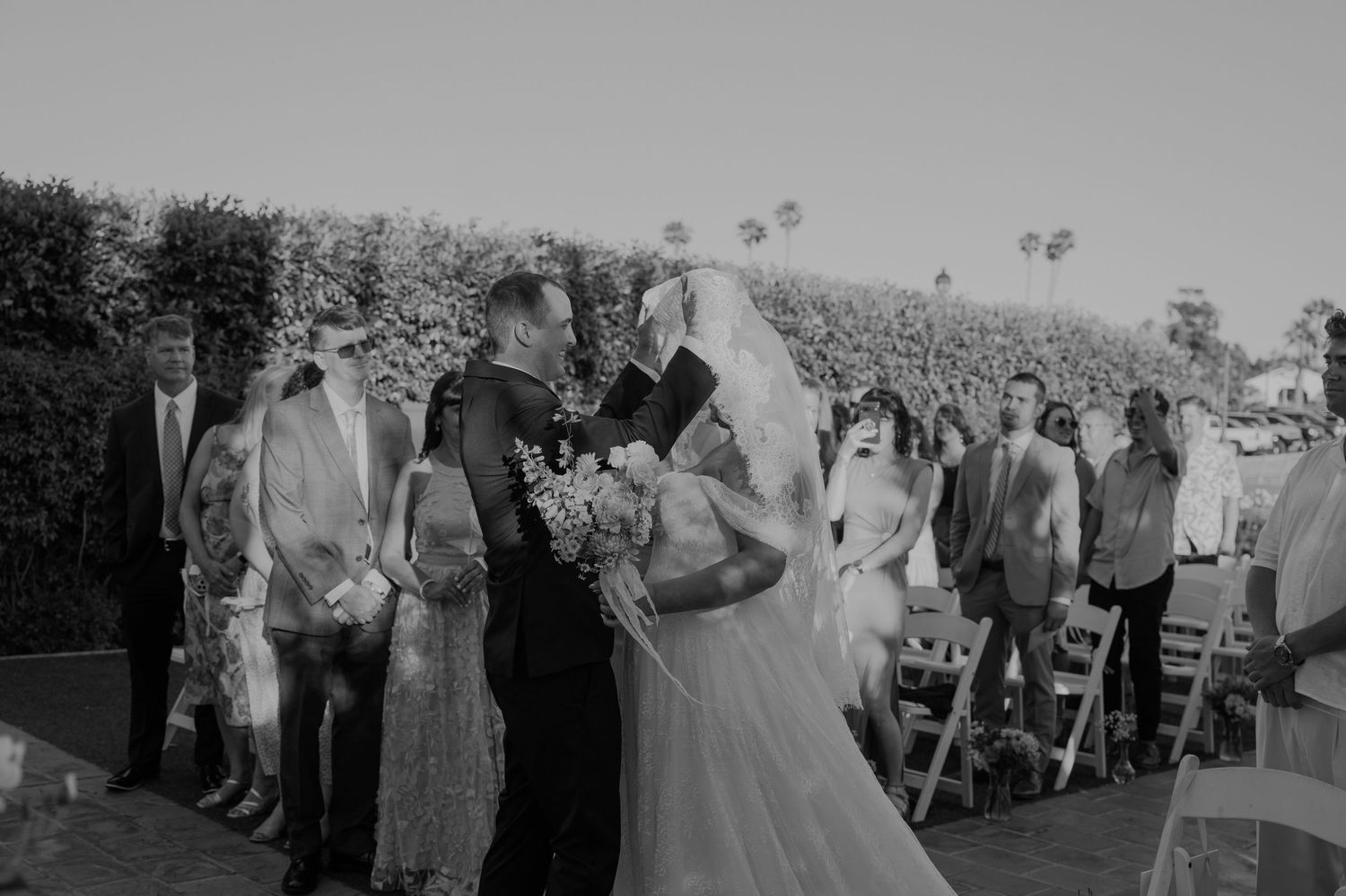 Black and white photo of a bride and groom sharing a kiss at an outdoor wedding ceremony with guests seated nearby.