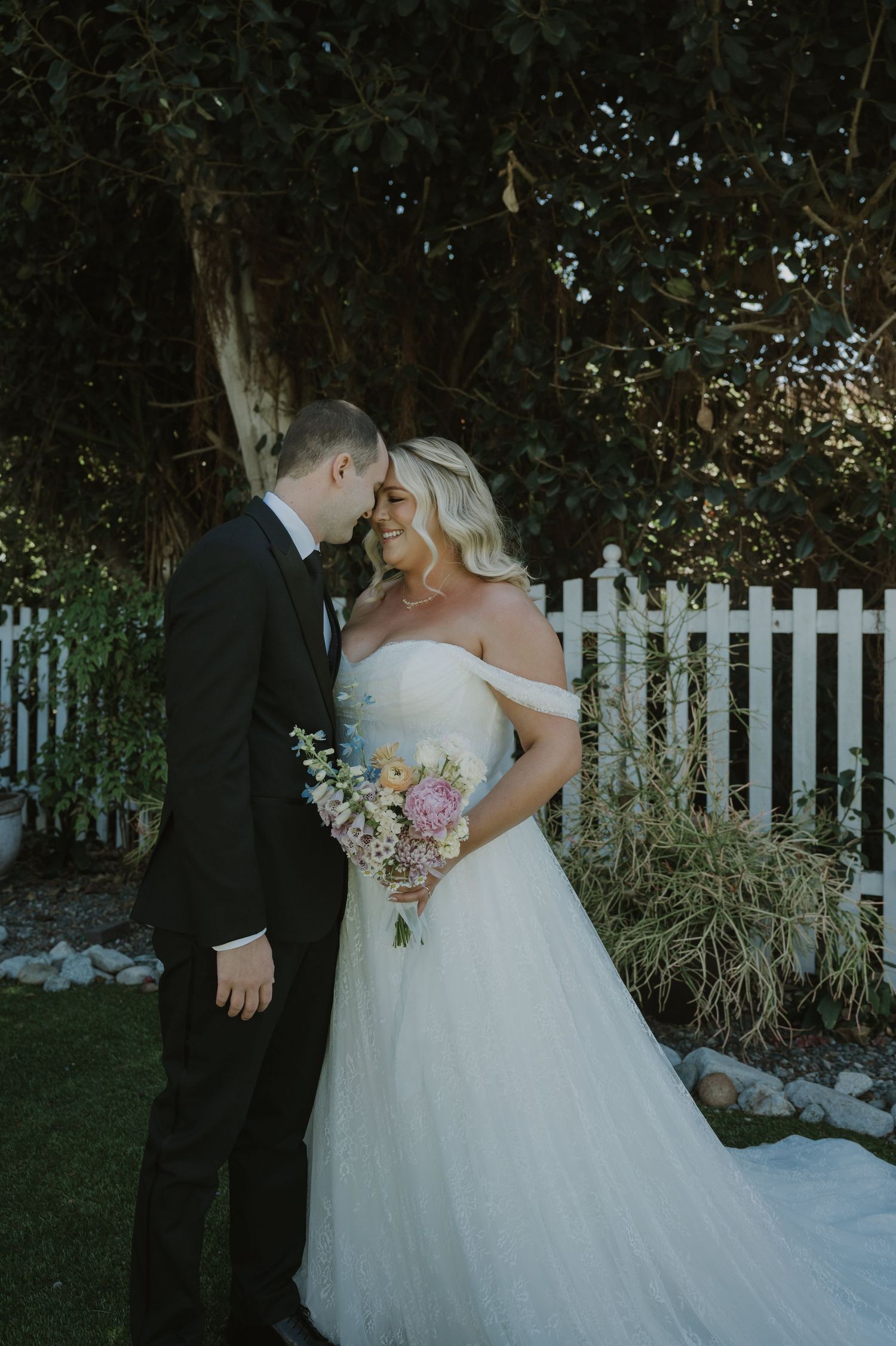 A bride in an off-shoulder white gown holds a colorful bouquet while sharing a romantic moment with her groom in a black tuxedo.