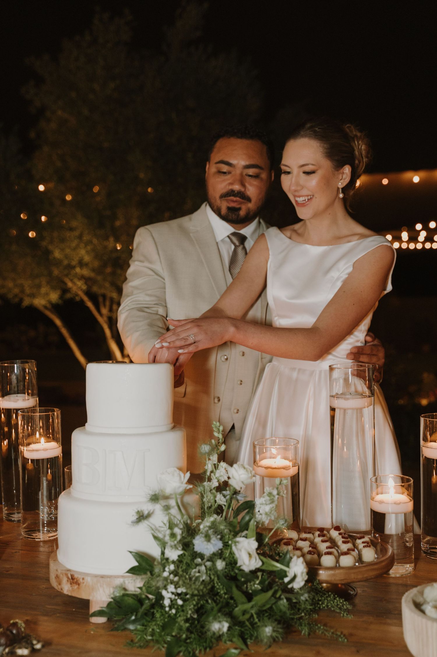 Bride and groom cutting elegant three-tier white wedding cake surrounded by candles at night.