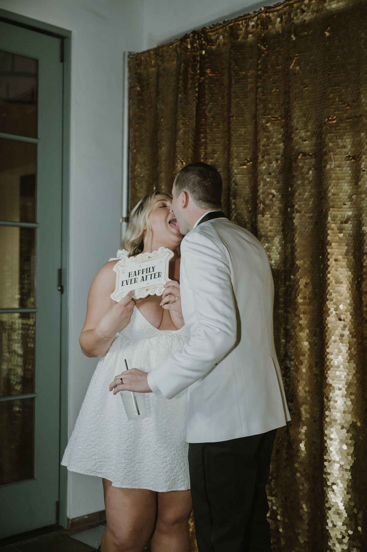 Bride and groom kissing at photo booth with gold sequin backdrop holding Happily Ever After sign.
