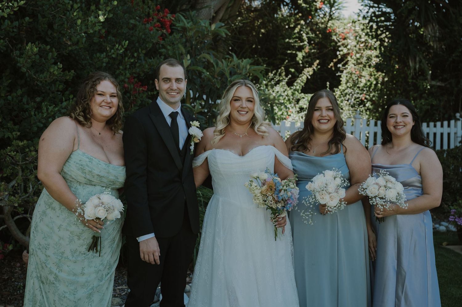 Bride in white gown with three bridesmaids in sage green dresses and groom in black suit posing outdoors.