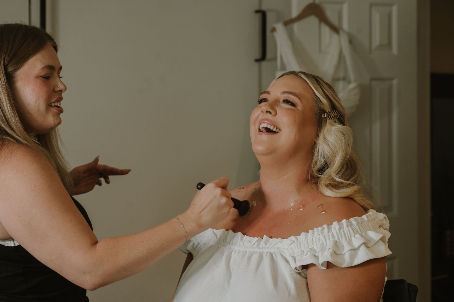 Bride laughing joyfully while bridesmaid applies makeup during wedding day preparations in a bright room.