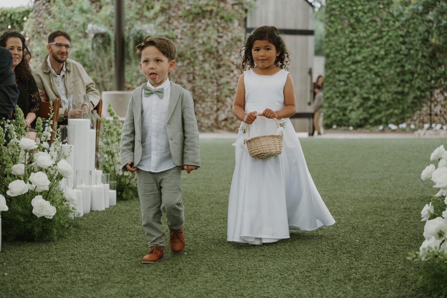 Ring bearer in gray suit and flower girl in white dress walking down wedding aisle lined with white floral arrangements.