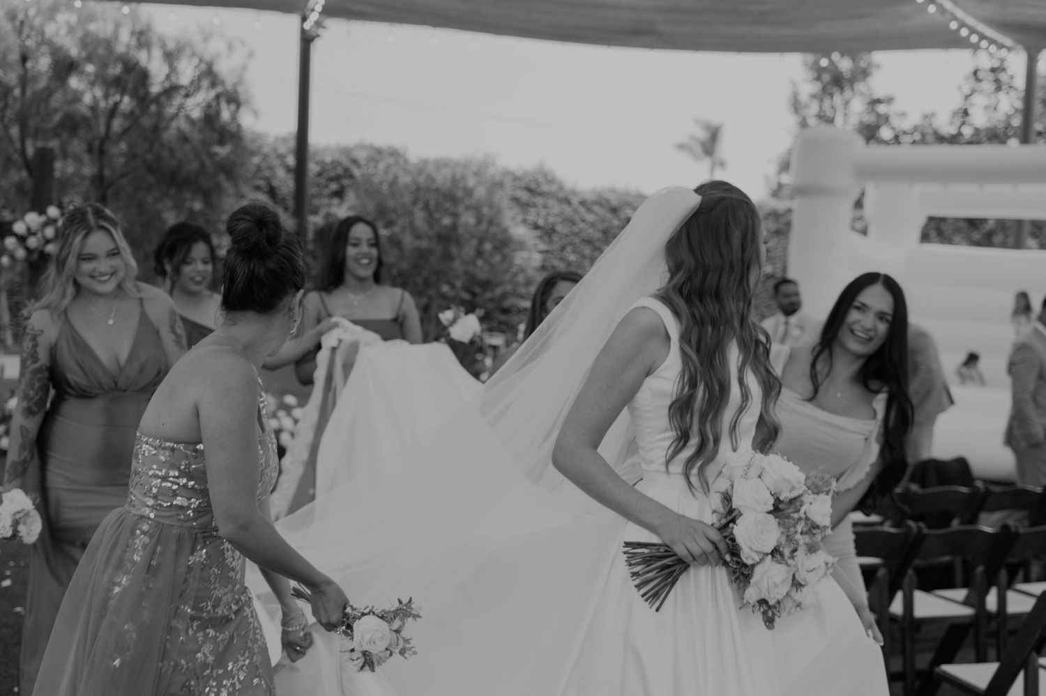 Bridesmaids helping bride with her white wedding gown and veil at outdoor ceremony.