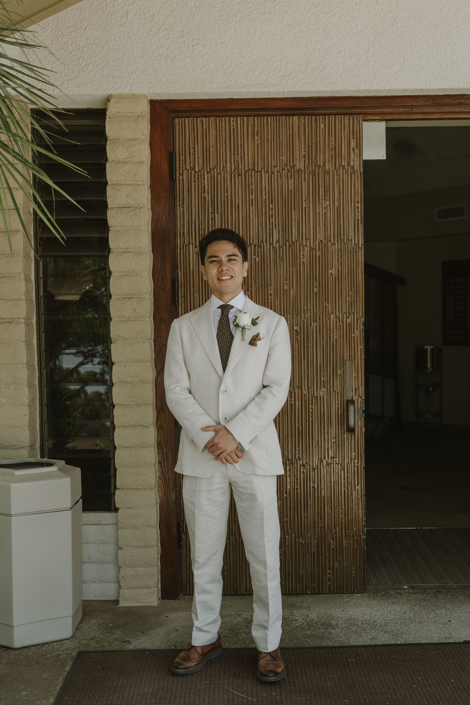 Groom in white linen suit with floral boutonniere standing before bamboo doors.
