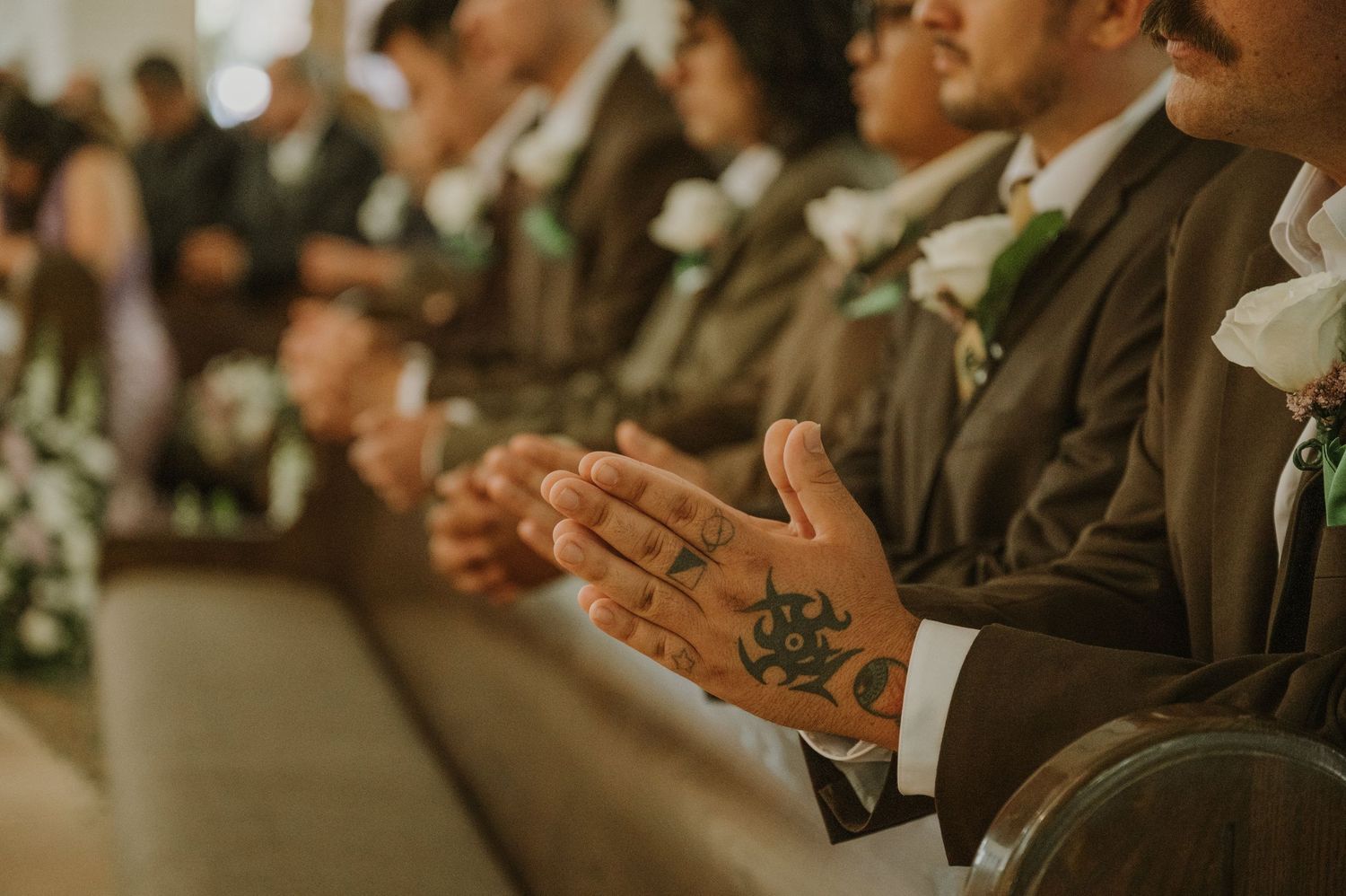 Close-up of groomsmen hands clasped in church pew, one hand featuring a decorative tattoo, during wedding ceremony.