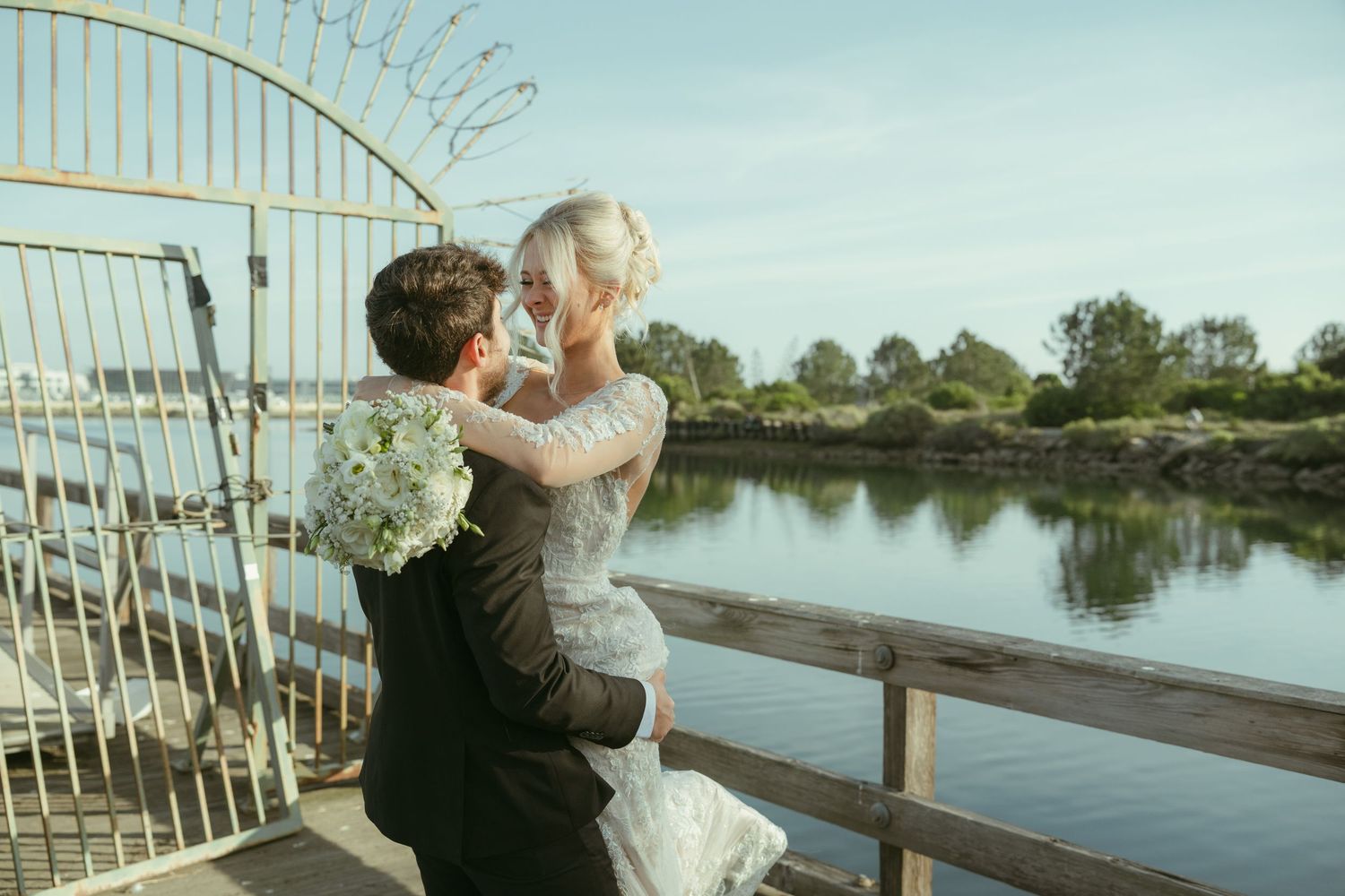 Bride and groom embrace on wooden dock by calm waterway, bride holding white hydrangea bouquet in romantic outdoor wedding photo.