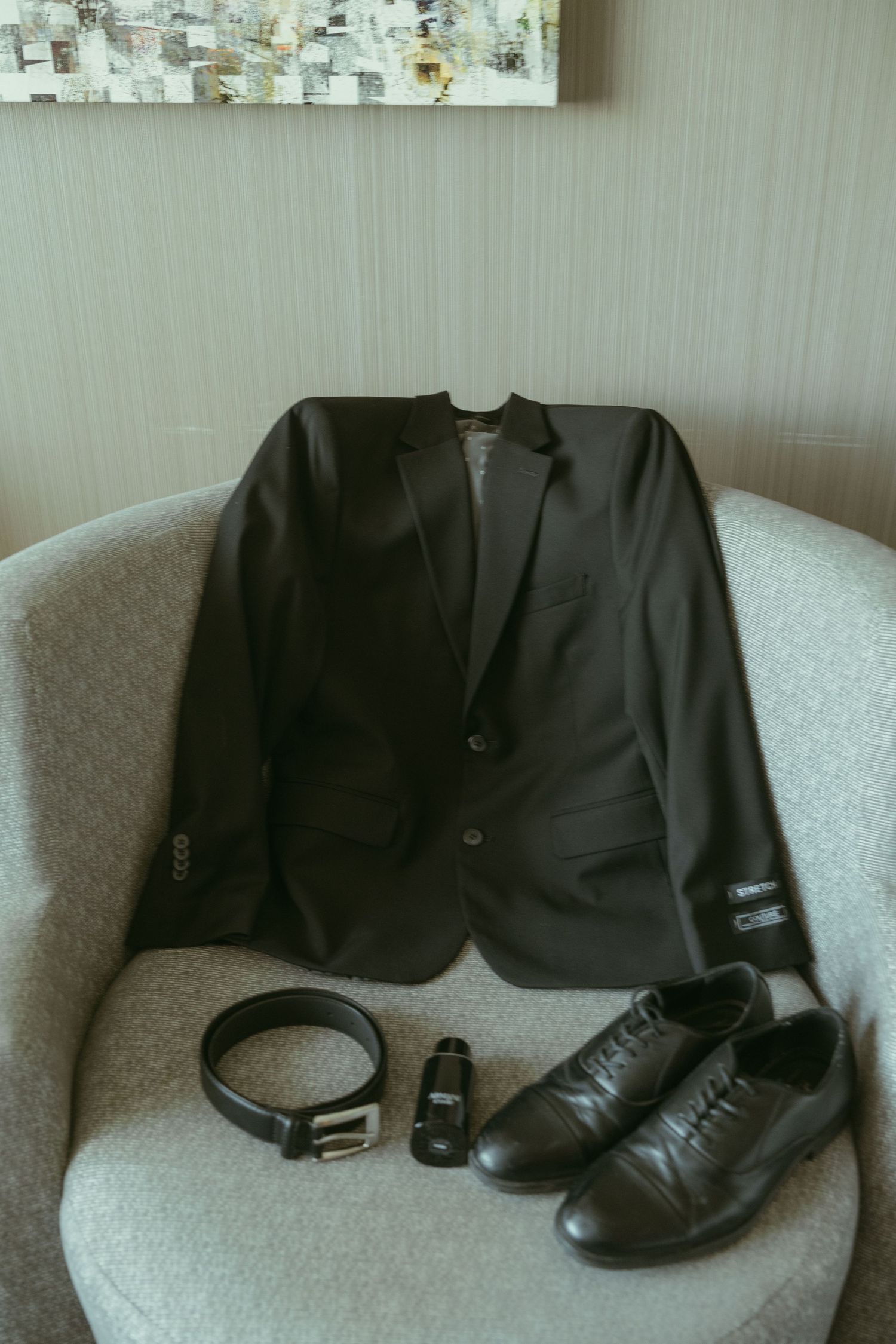 Black suit jacket, leather belt, cologne, and dress shoes arranged on a gray chair for a groom preparation.