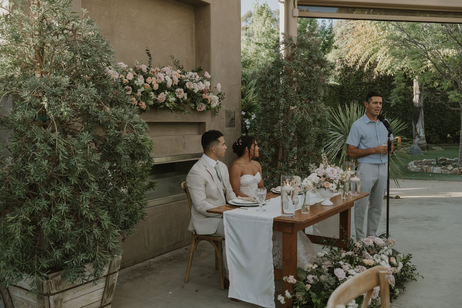A couple sits at a floral-decorated sweetheart table while a guest delivers a wedding speech.