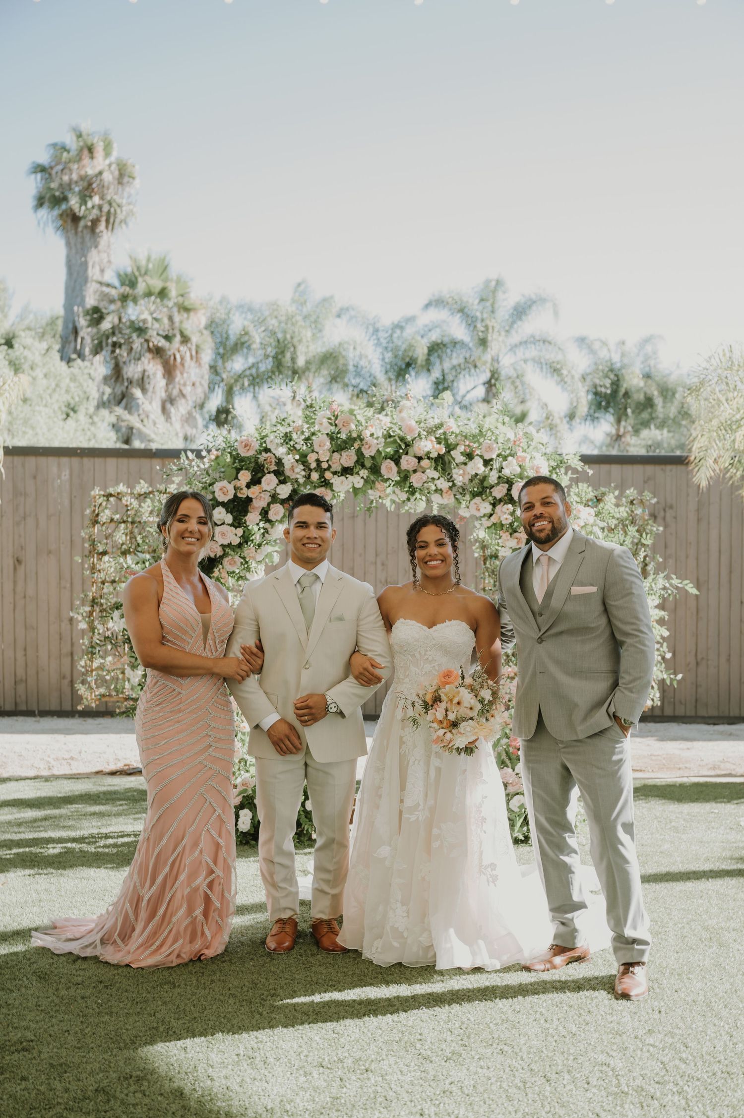 Wedding party of four posing outdoors with floral arch, bride in white gown, bridesmaid in pink, groomsmen in gray suits.
