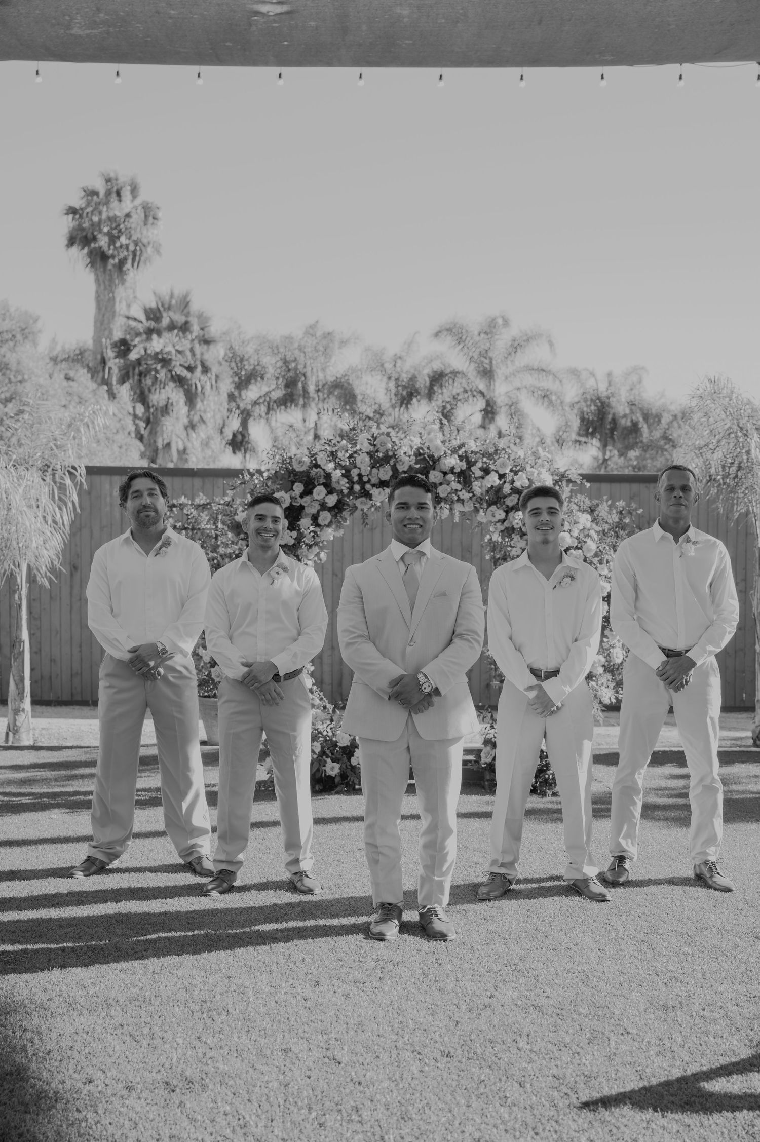 Black and white photo of groom and four groomsmen standing formally in front of a floral arch outdoors.