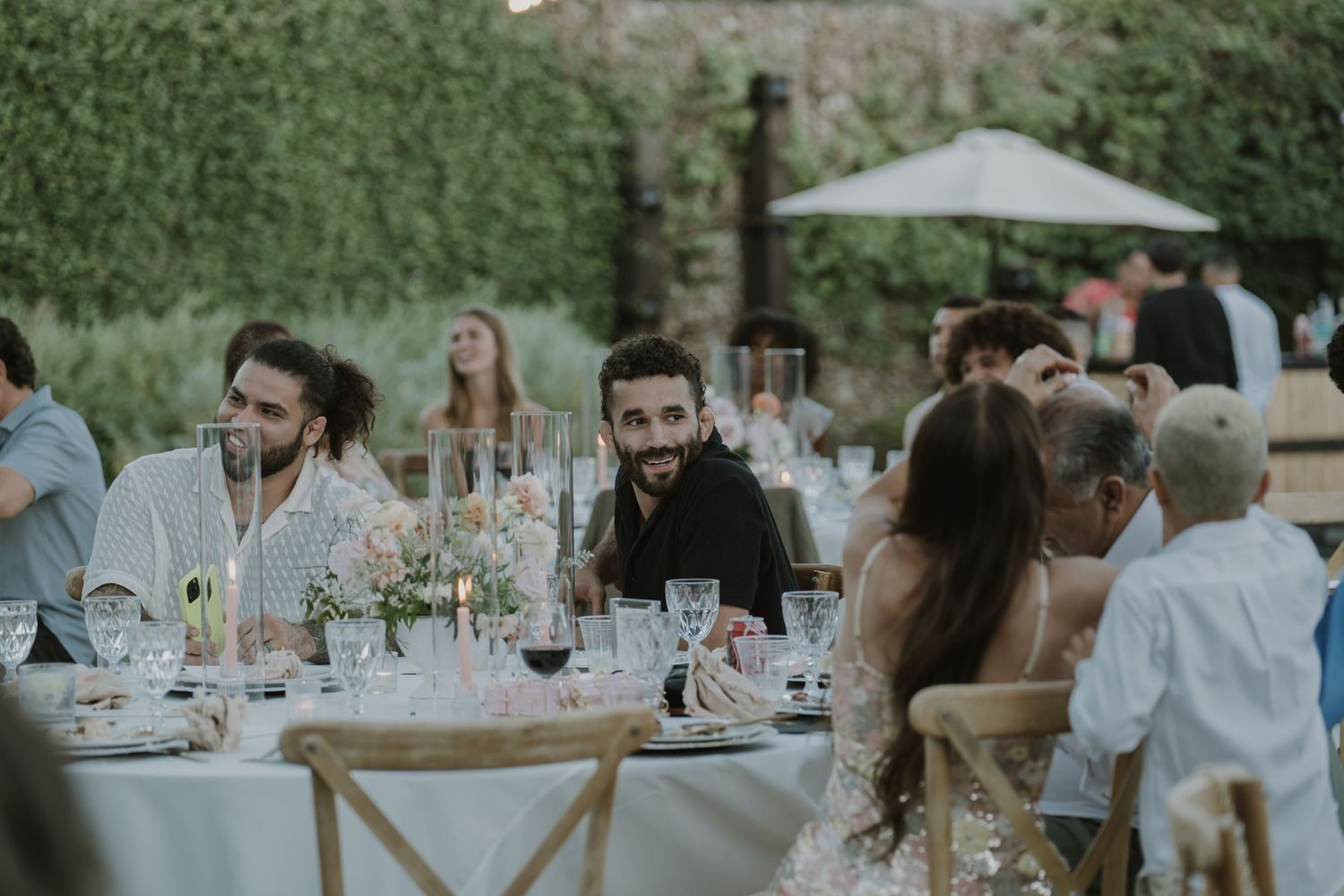 Guests smiling and laughing at elegantly set outdoor wedding reception tables surrounded by lush greenery.