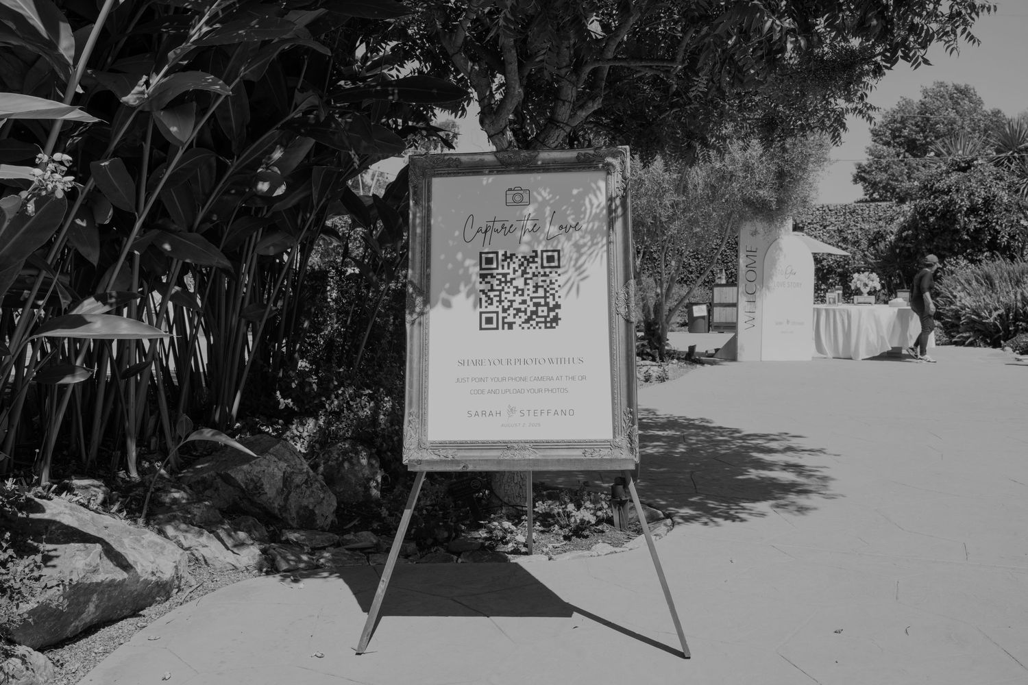 Black and white photo of a wedding welcome sign with QR code on easel in outdoor garden venue.