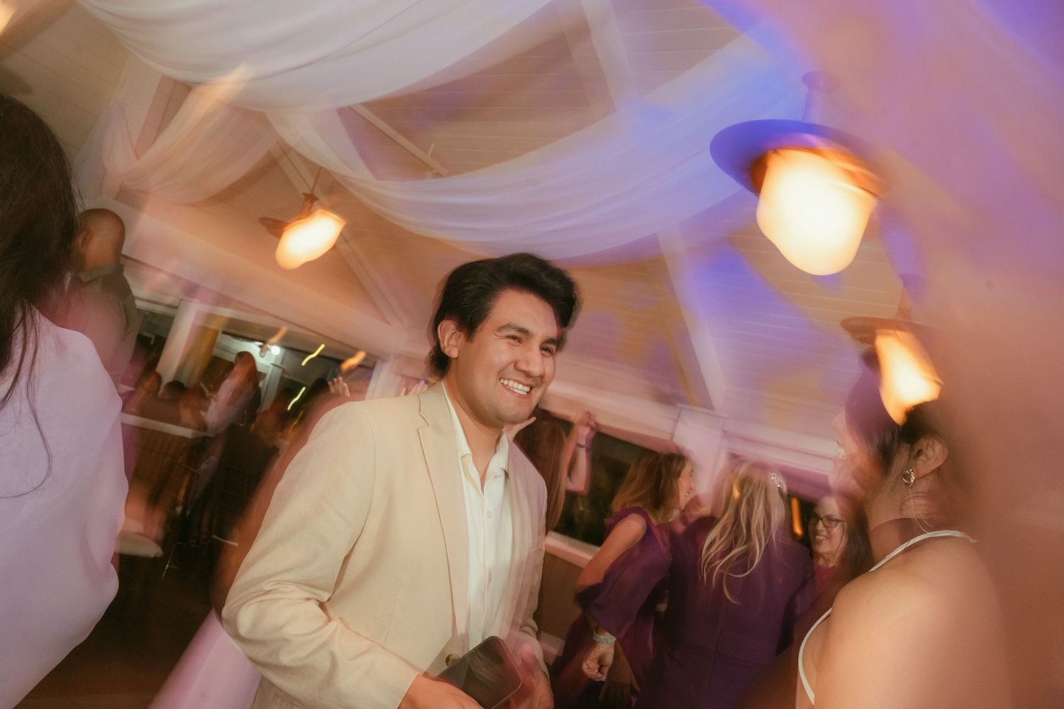 Smiling man in cream blazer dancing at elegant wedding reception with draped ceiling and warm lighting.