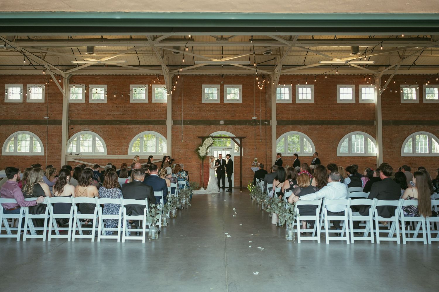 Wide-angle view of indoor wedding ceremony in historic brick hall with white chairs and decorative arch at altar.