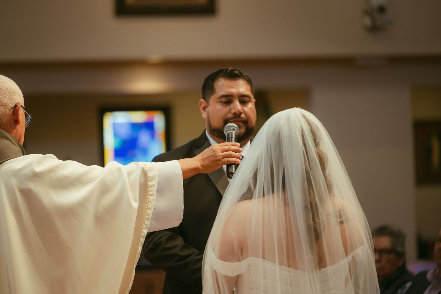 Groom speaks into microphone during wedding ceremony at church altar with bride in veil and officiant in white robe.