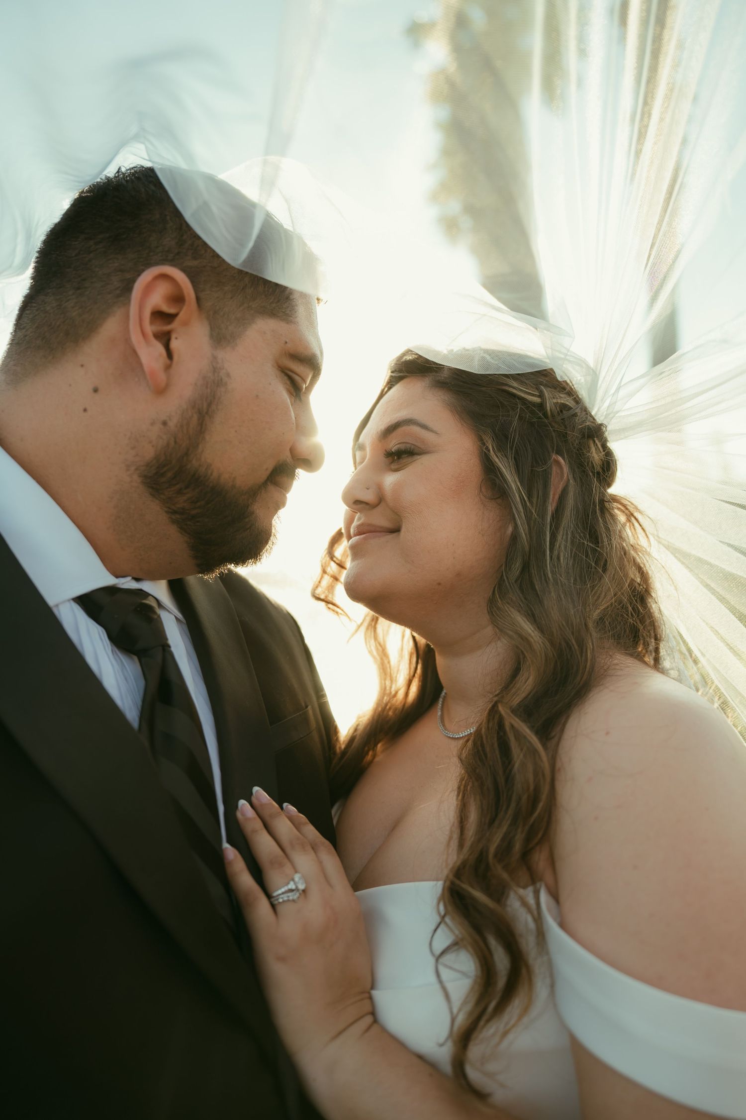 Bride and groom share an intimate moment under a veil, with the bride smiling and wearing an off-shoulder gown.