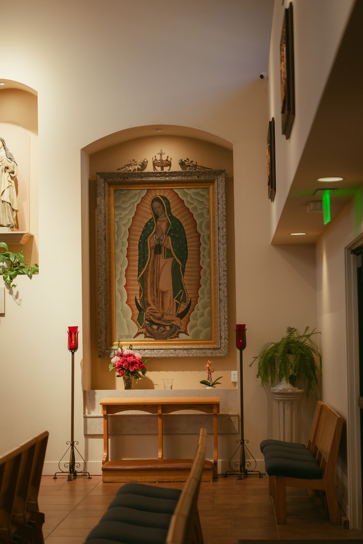 Catholic chapel altar with Our Lady of Guadalupe painting, candles, and fresh flowers in a warmly lit niche.