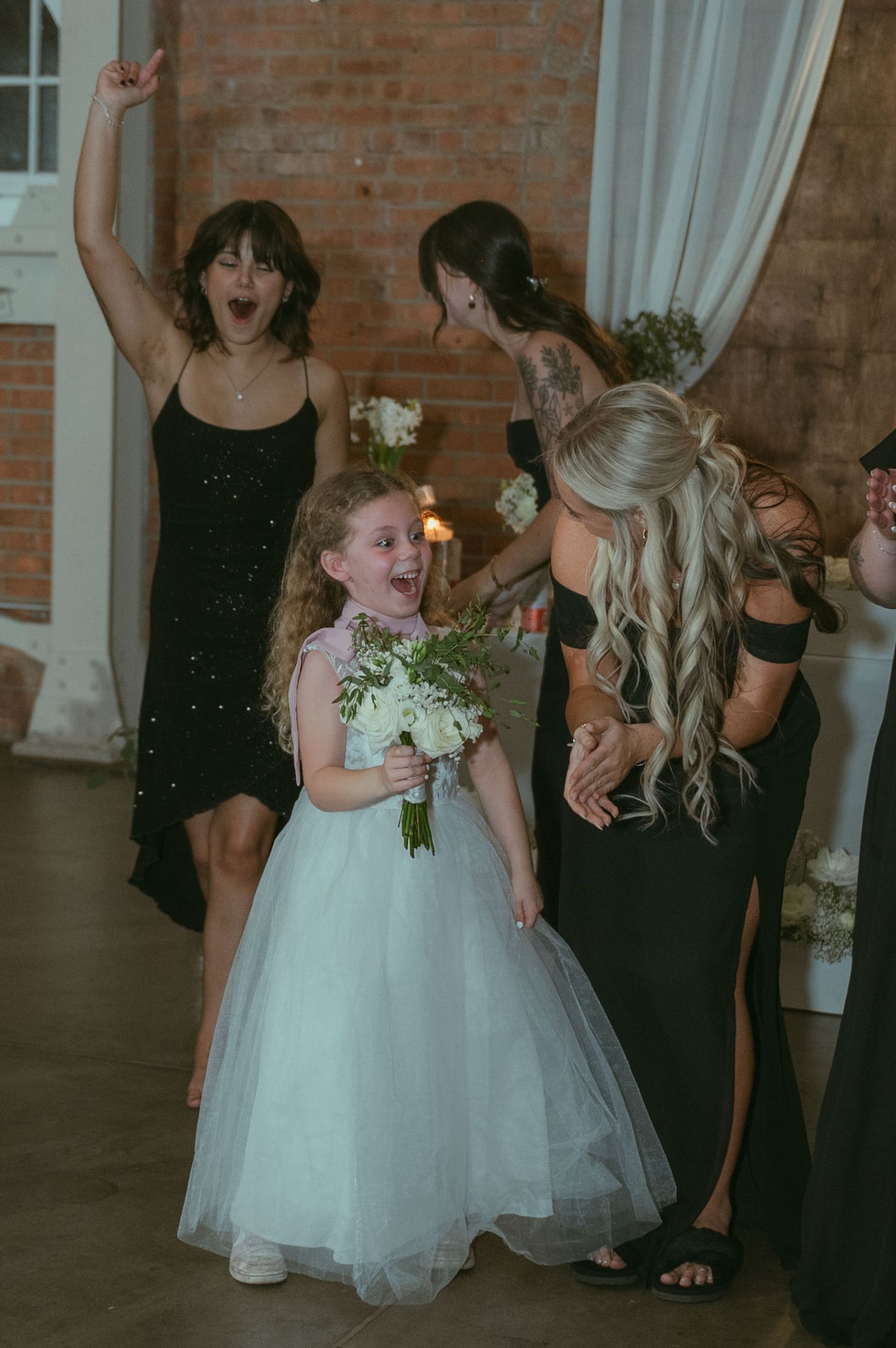Excited flower girl in white tulle dress holds bouquet surrounded by bridesmaids in black dresses at wedding.