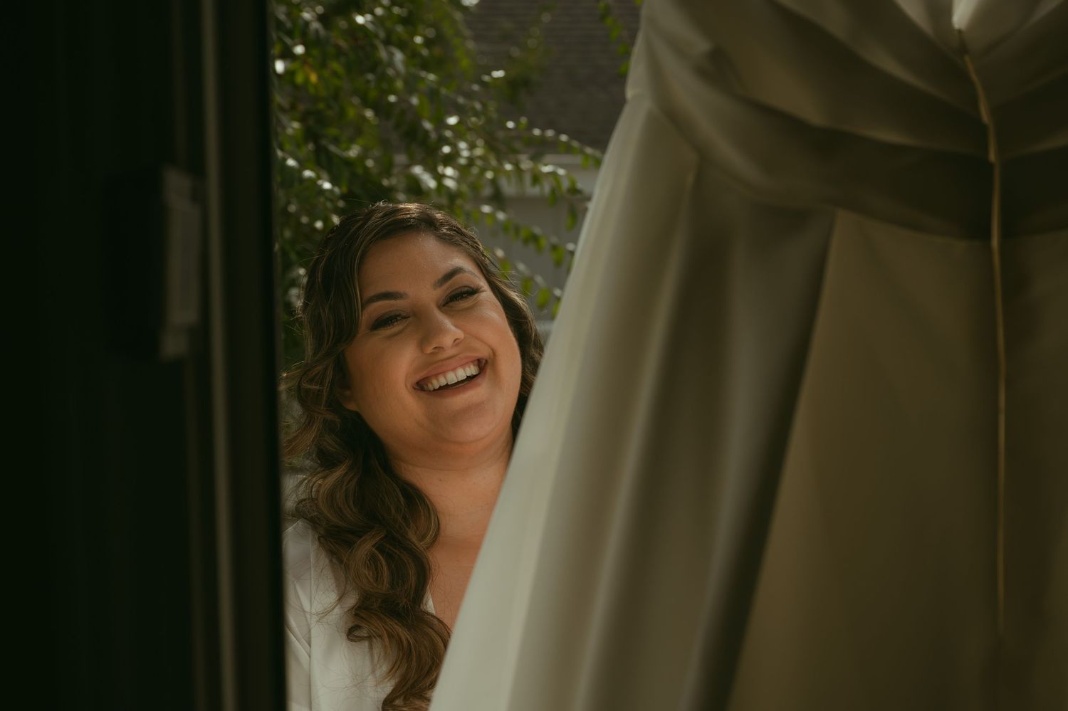 Smiling bride in white robe peeks through flowing curtain near window on wedding day.