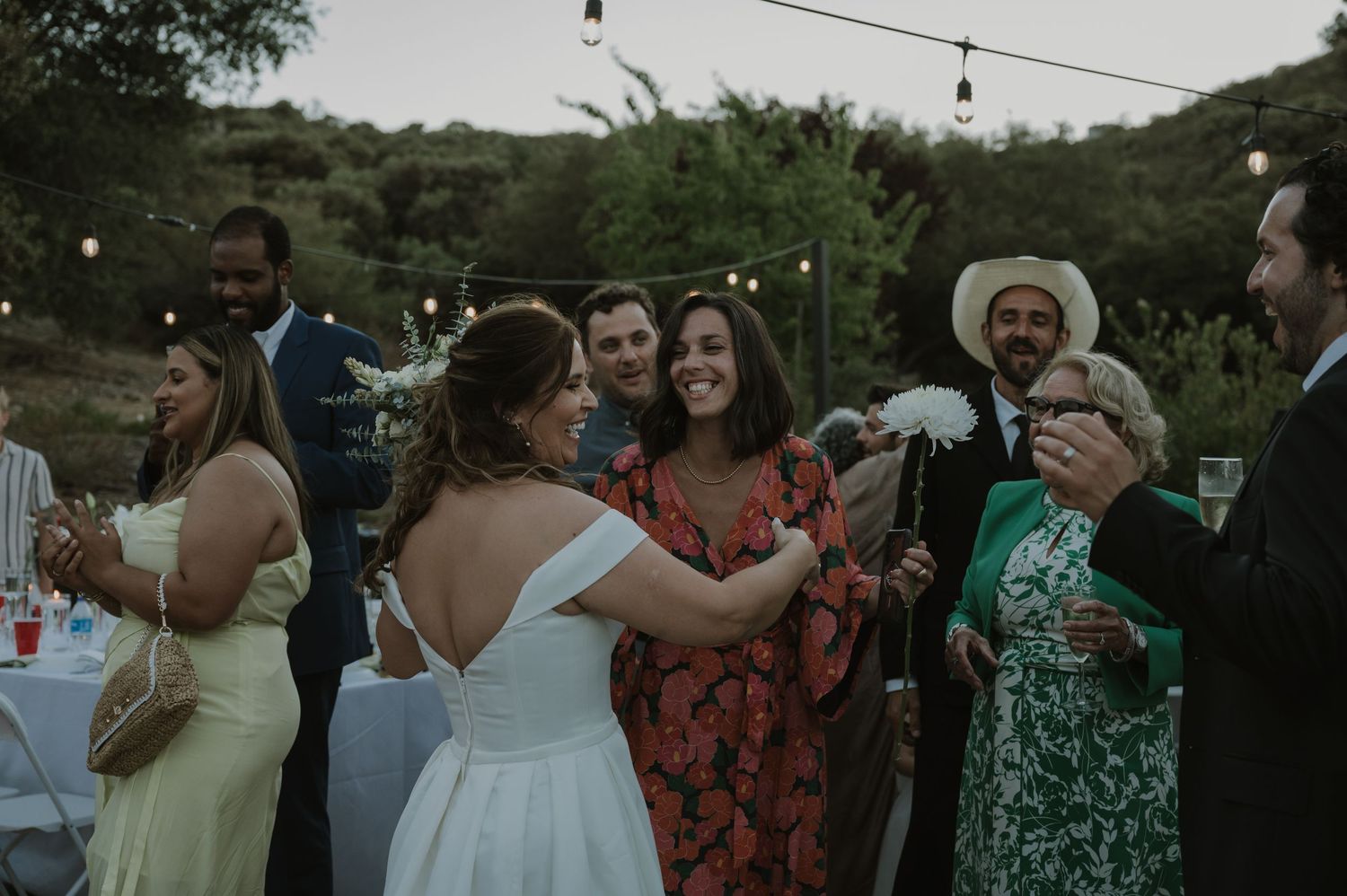 Bride dancing with guests at an outdoor wedding reception under string lights at dusk.