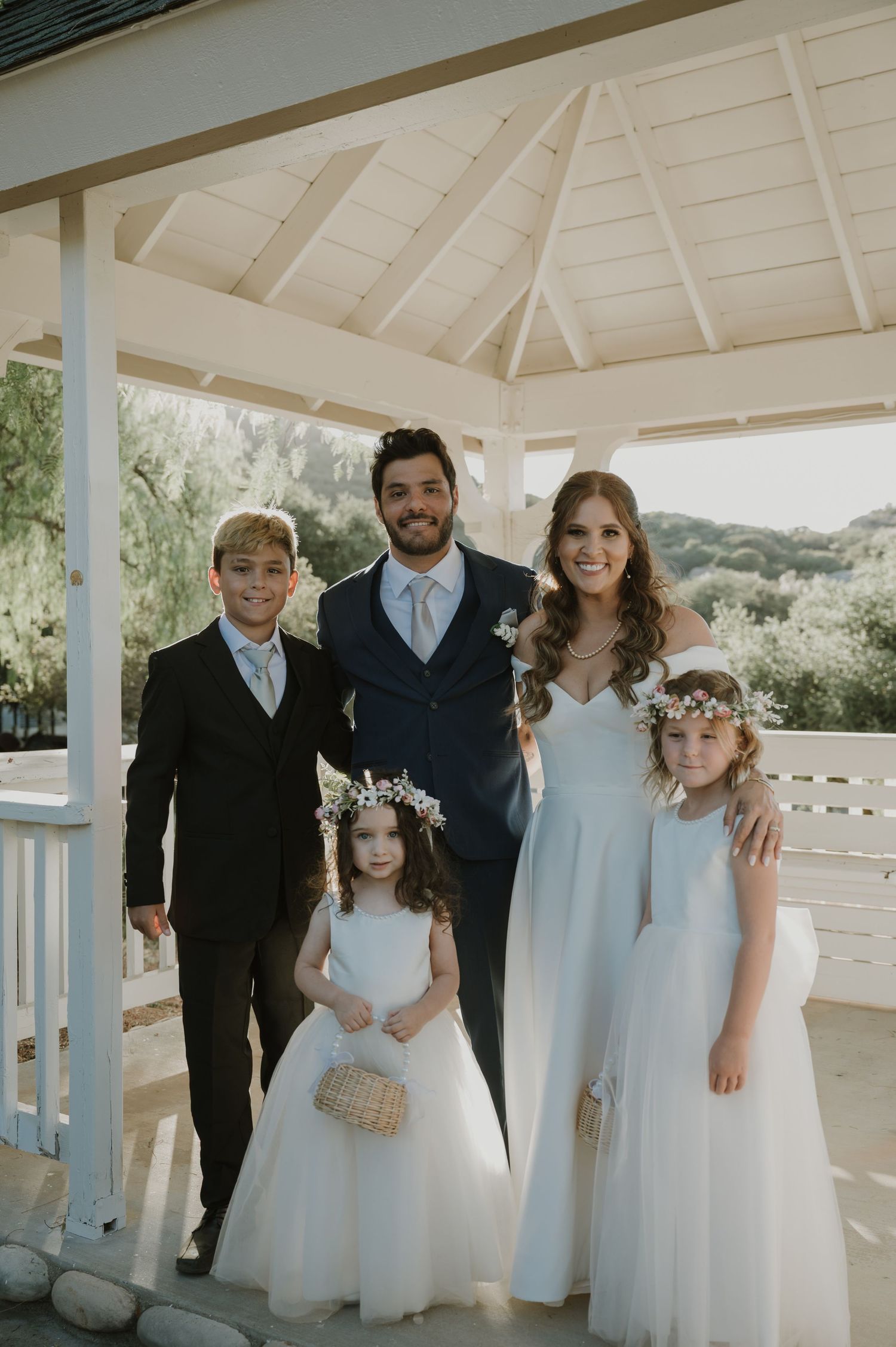 Bride and groom posing with three children in white dresses and suit under wedding gazebo.