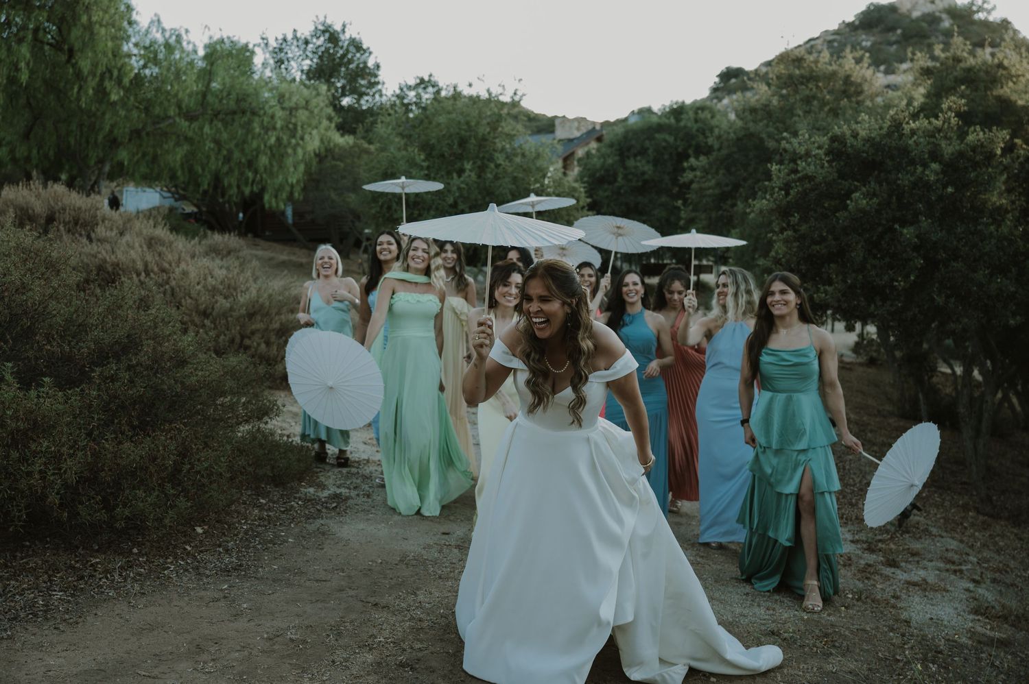 Bride leading bridesmaids in colorful dresses along outdoor path at wedding celebration.