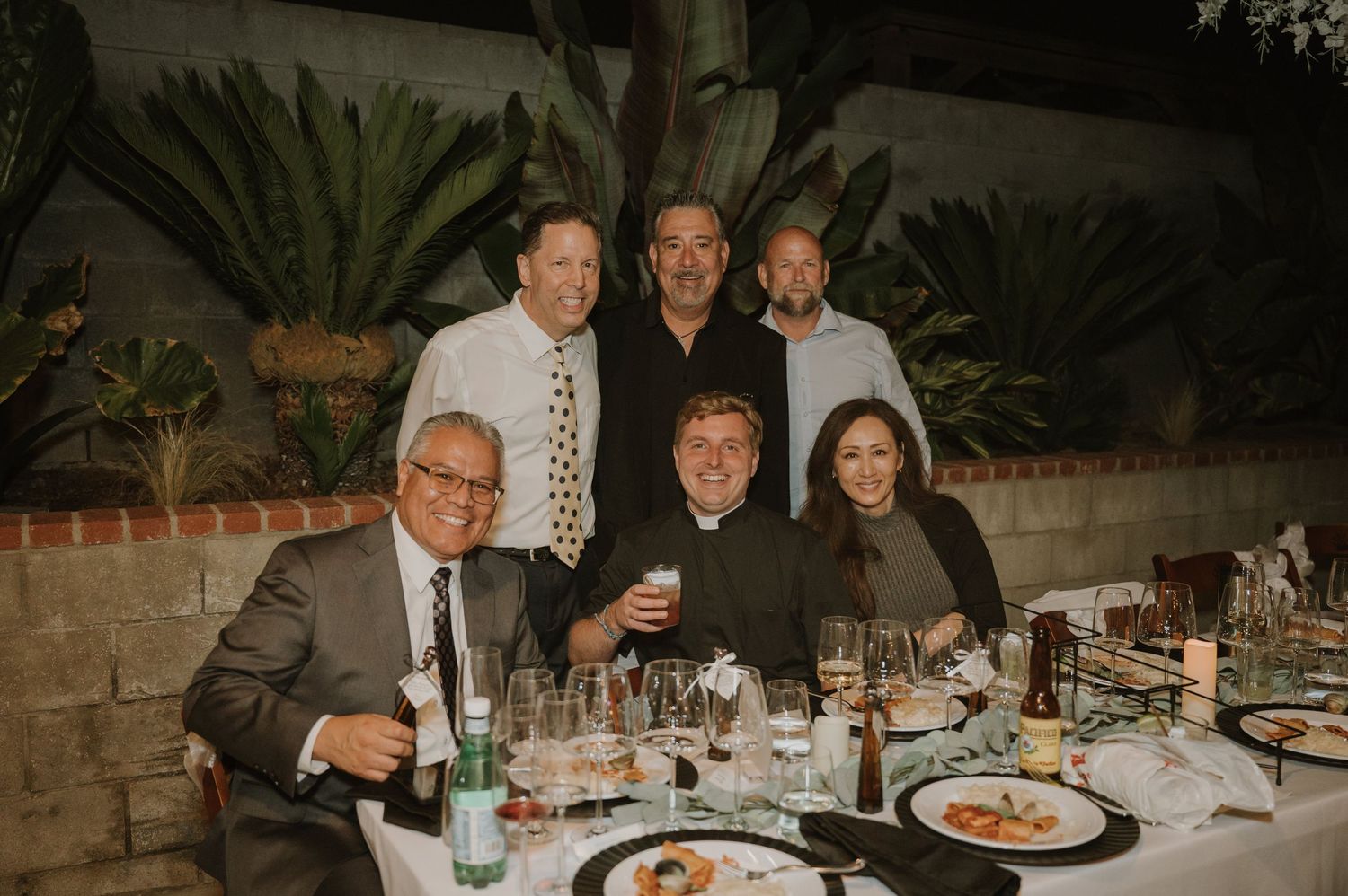 Six people pose and smile at a formal dinner table with wine glasses and plates in an outdoor evening setting.