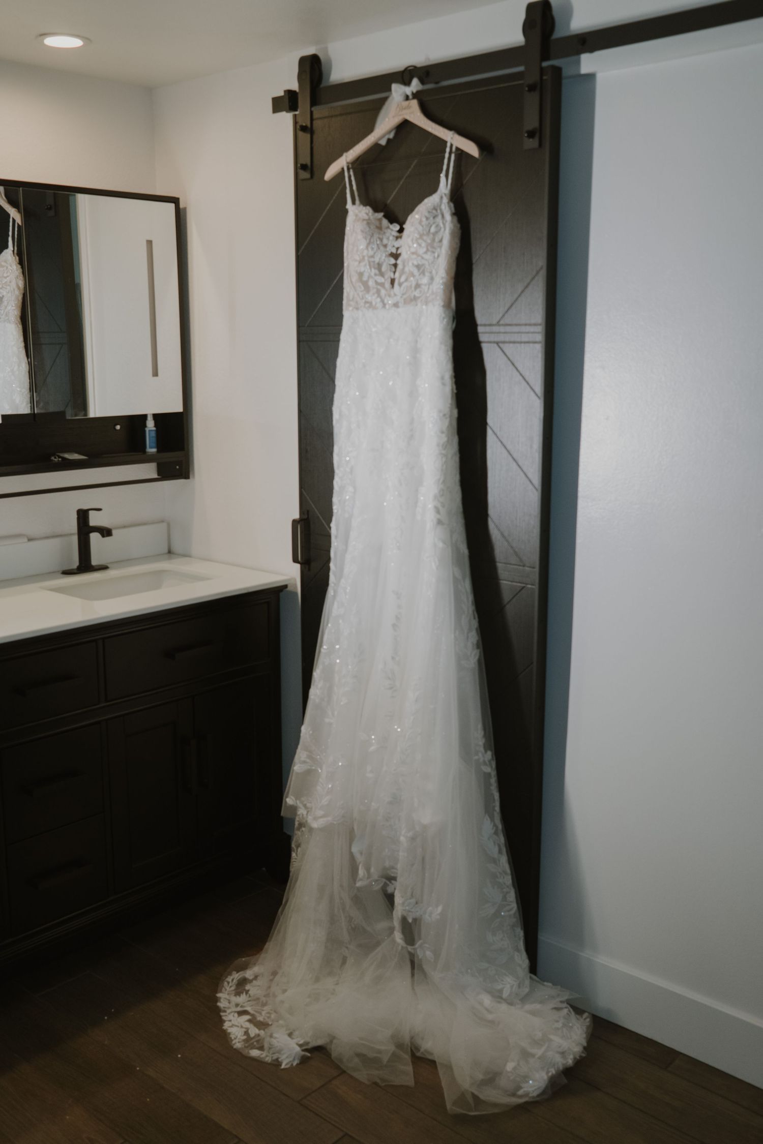 White lace wedding dress with spaghetti straps hanging on a barn door in a modern bathroom setting.