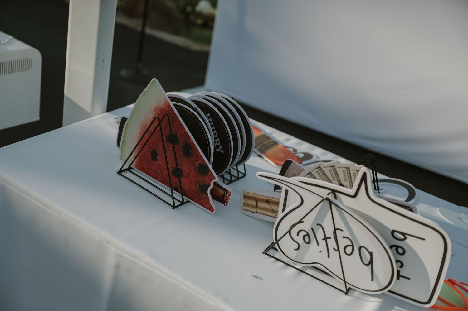 Decorative watermelon-shaped fans and best besties sign displayed on white tablecloth at outdoor party.