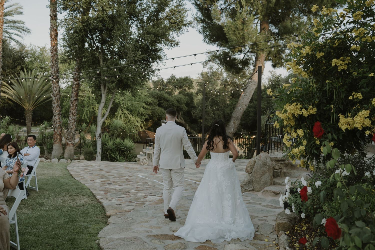 Bride and groom walk hand in hand down garden wedding aisle surrounded by lush greenery.