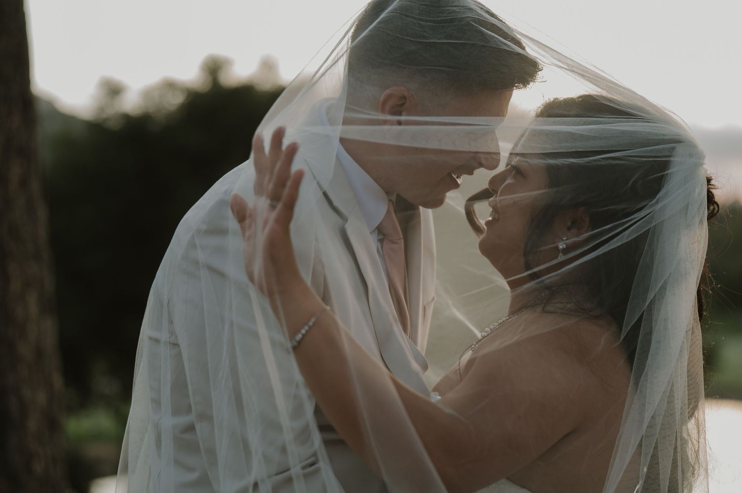 Bride and groom share an intimate moment under a flowing wedding veil at golden hour.