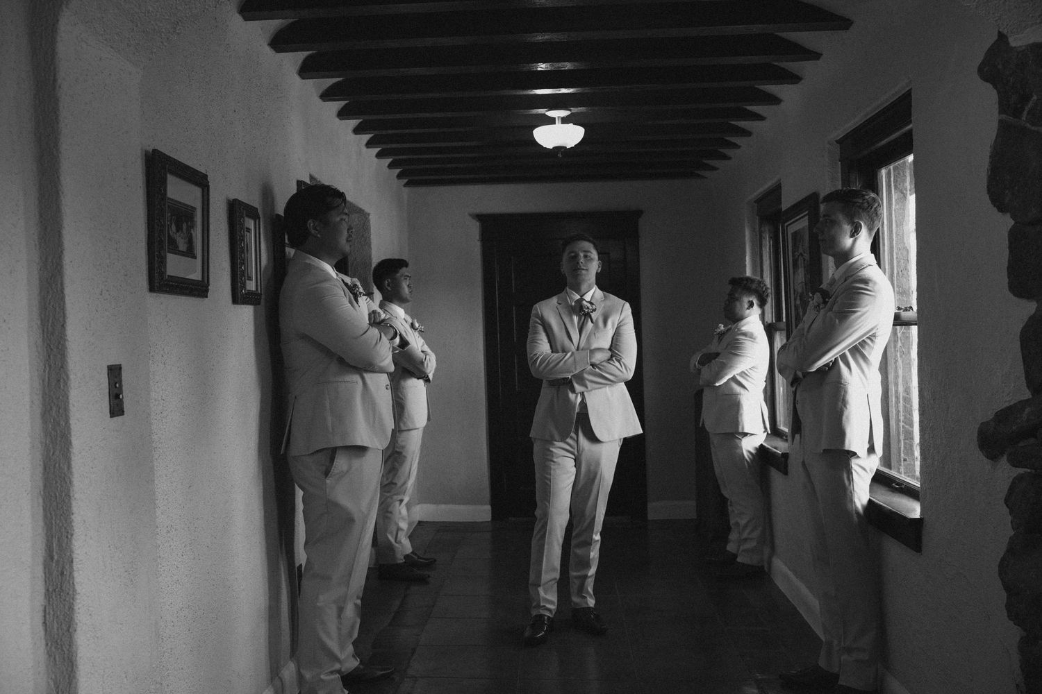 Groomsmen in light suits wait in a hallway in a black and white wedding photo.