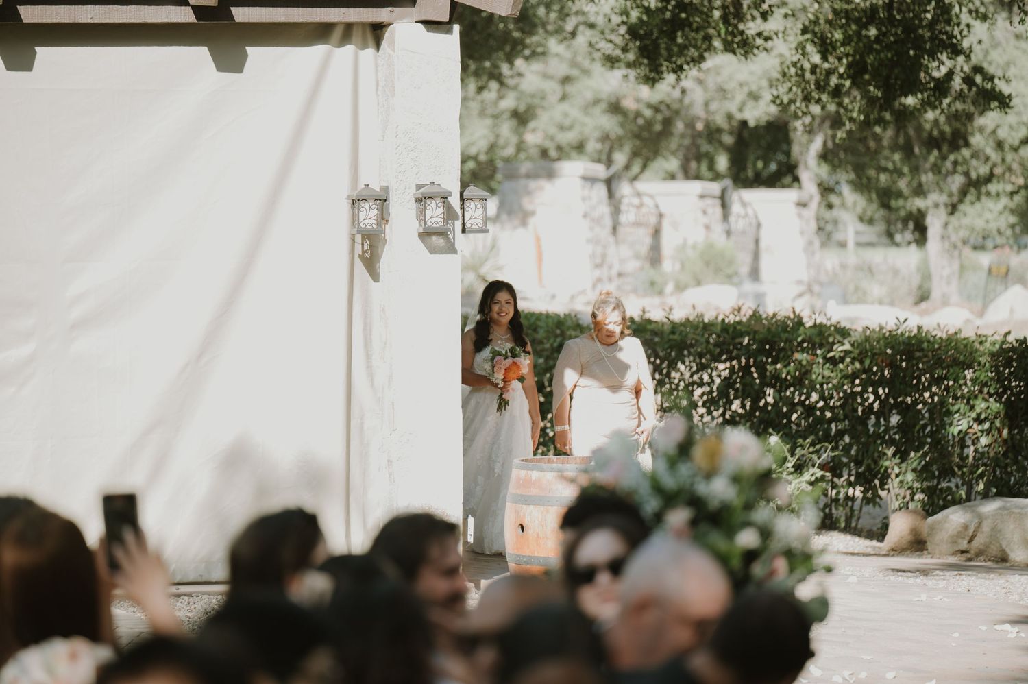 Bride and bridesmaid walk down outdoor wedding aisle surrounded by lush greenery and white tent.