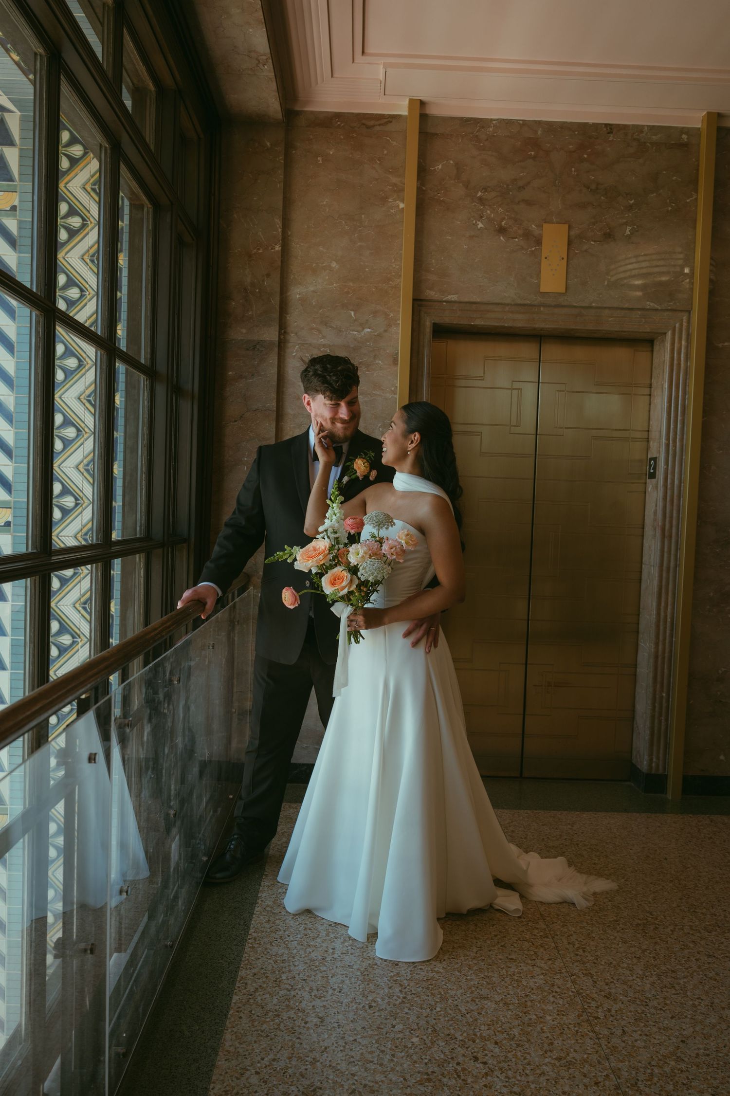 Bride and groom sharing intimate moment indoors, bride holding colorful bouquet, groom in dark suit.