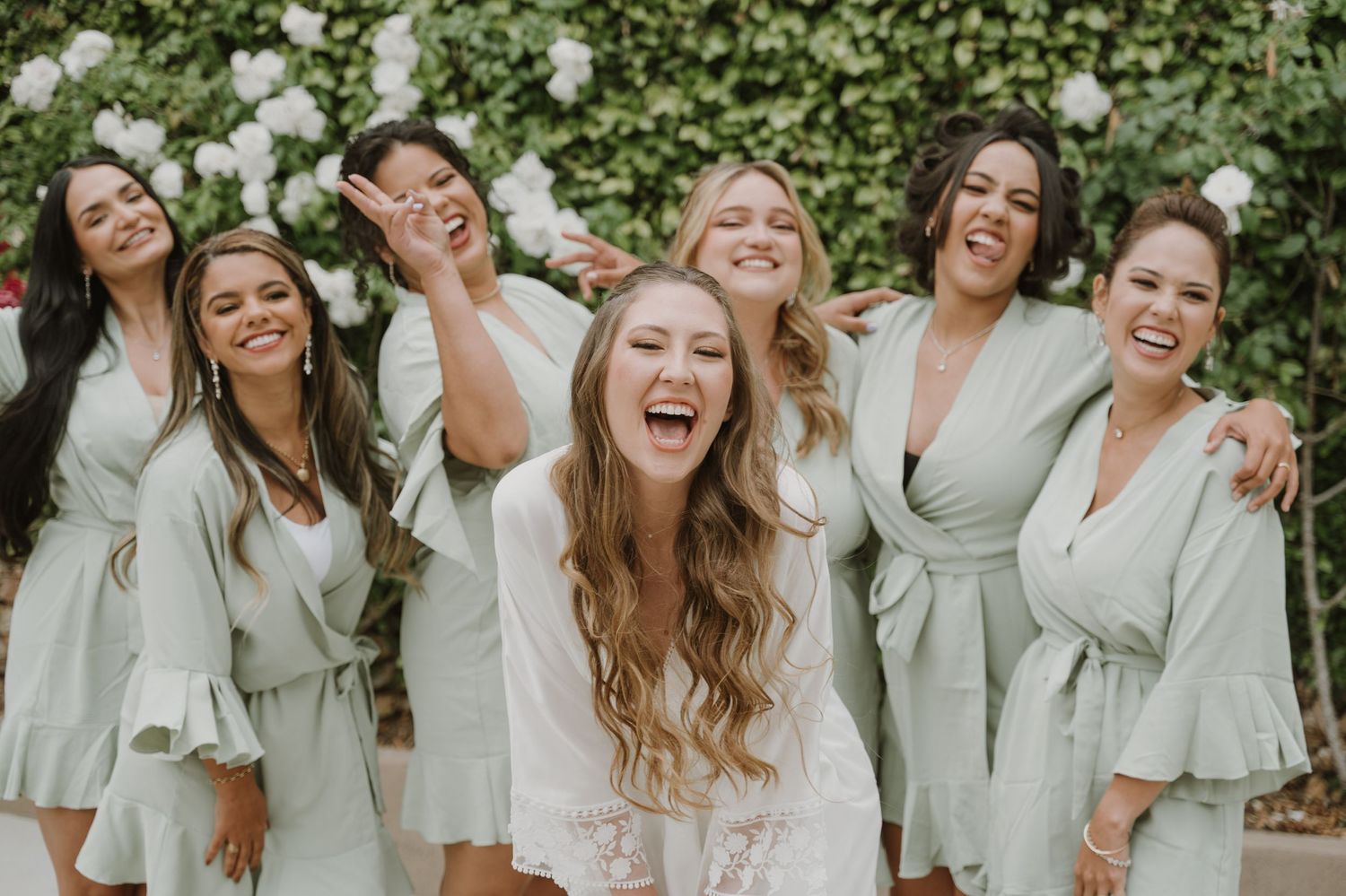 Bride laughing with bridesmaids in sage green robes posing outdoors in front of lush white floral greenery.