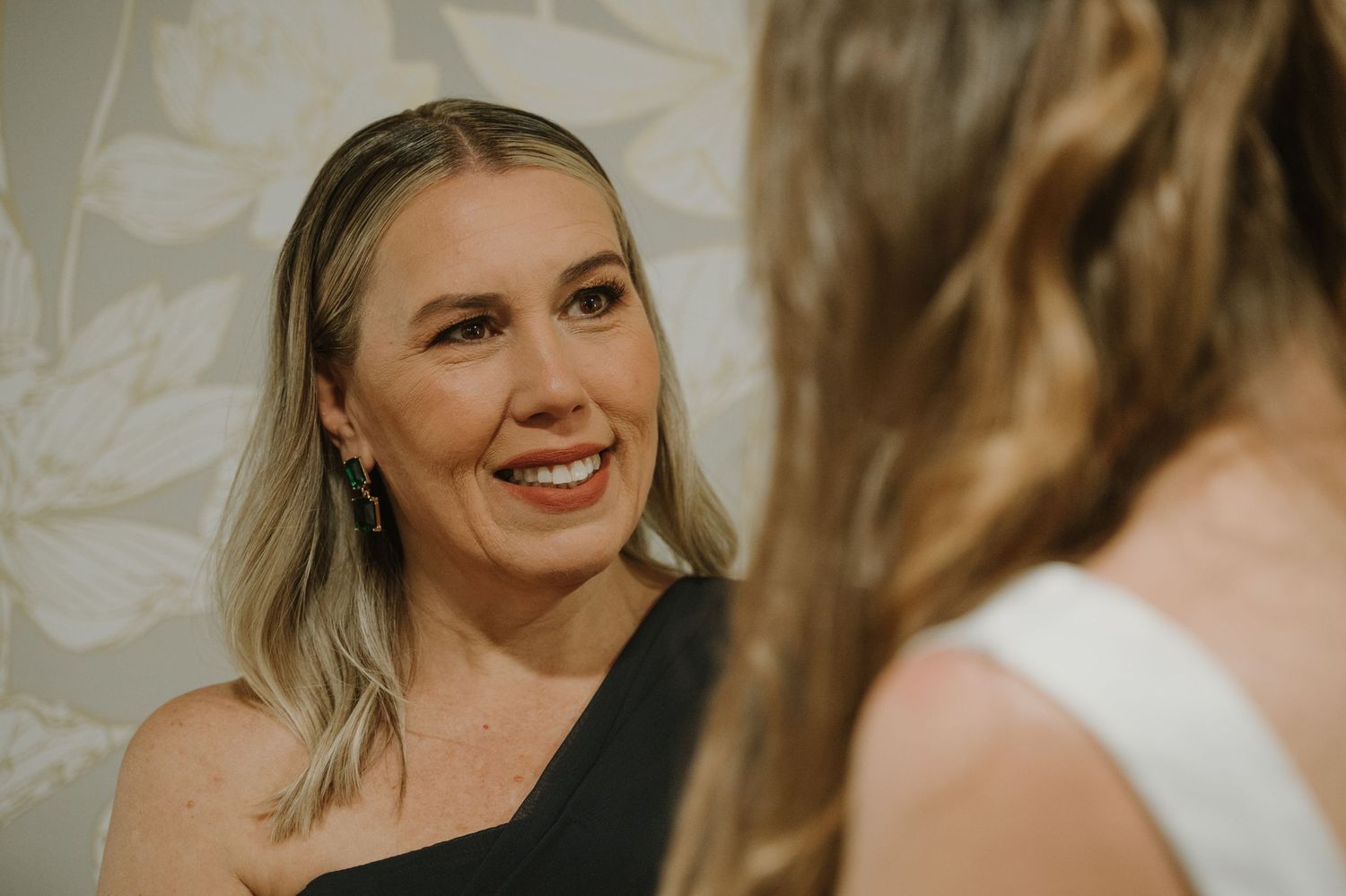 Smiling blonde woman in black one-shoulder dress looking warmly at bride during wedding preparations.