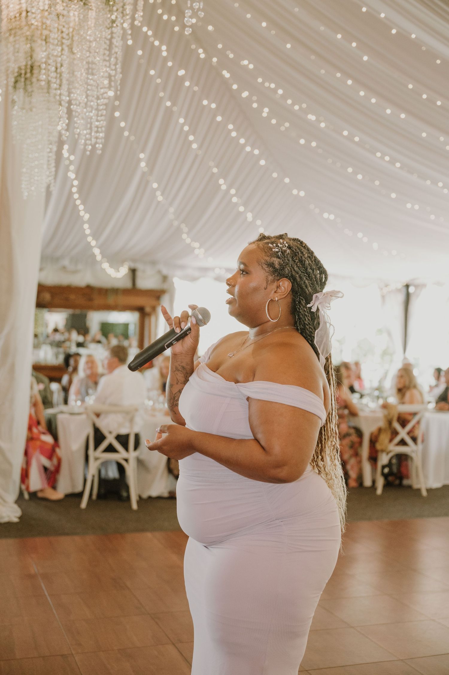 Woman in white off-shoulder dress singing into microphone at elegant wedding reception under draped lights.