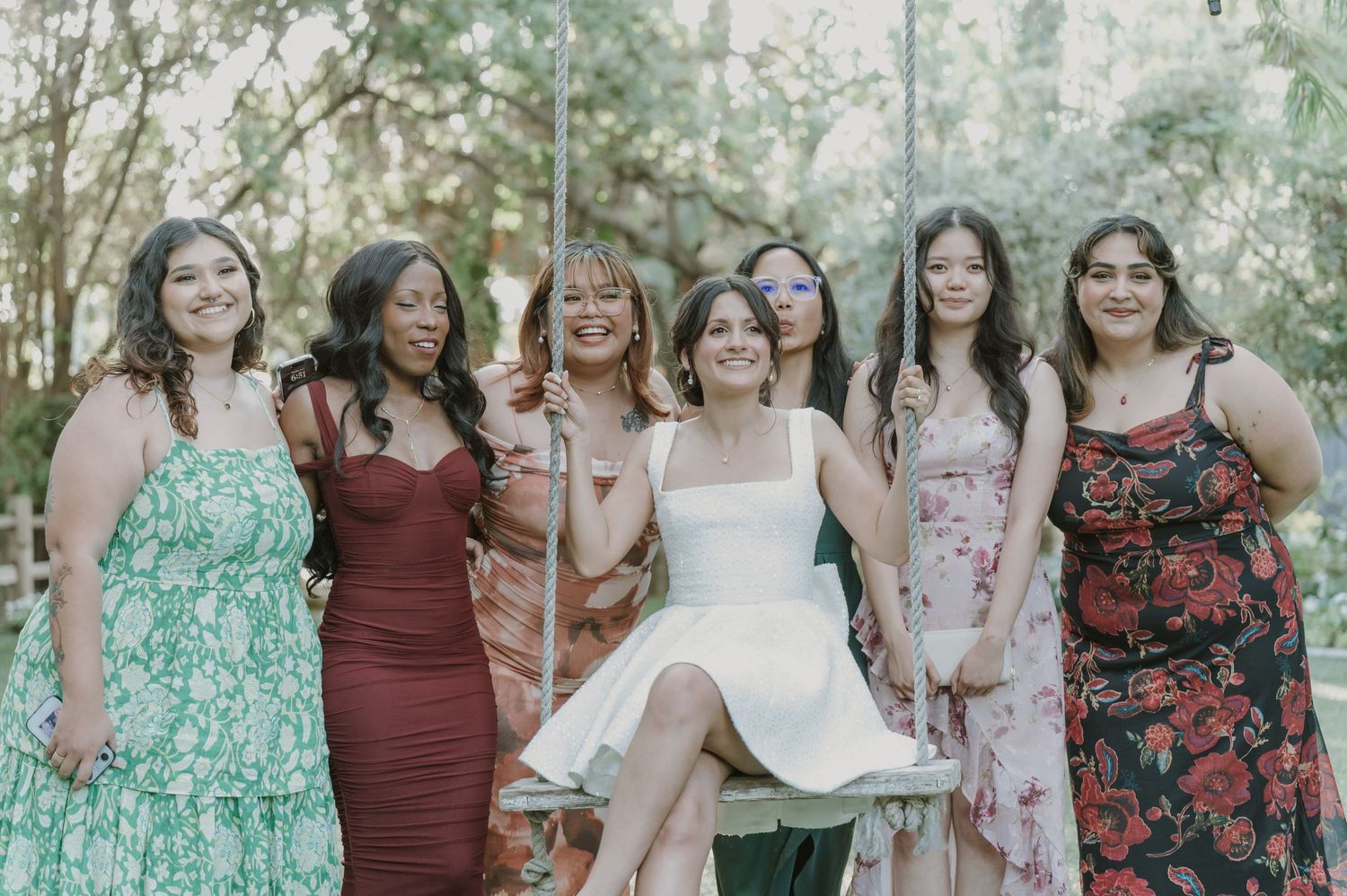 Bride in white dress posing with six friends in colorful gowns outdoors at wedding celebration.