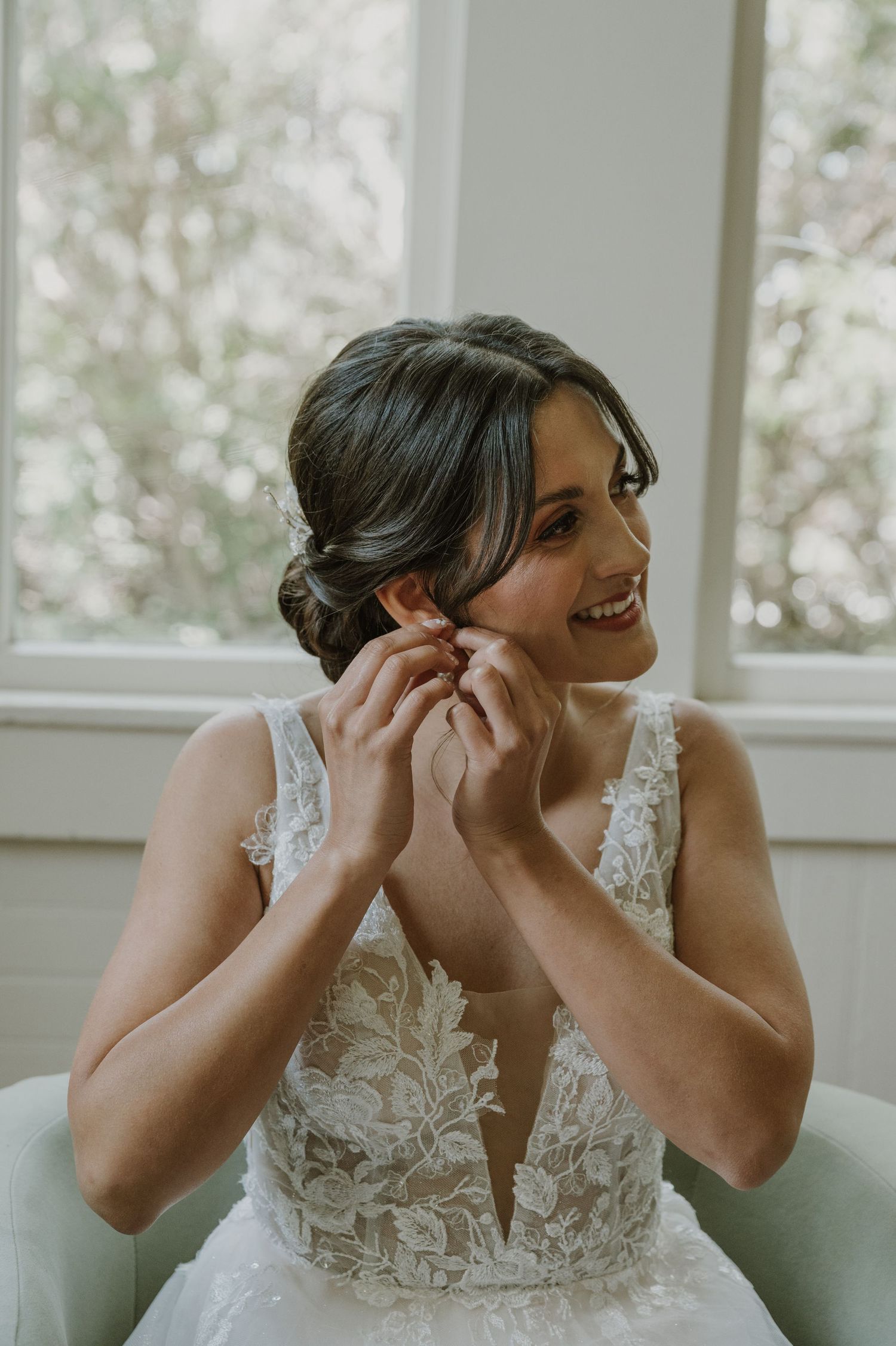 Smiling bride in lace wedding gown putting on pearl earring while seated near bright window.