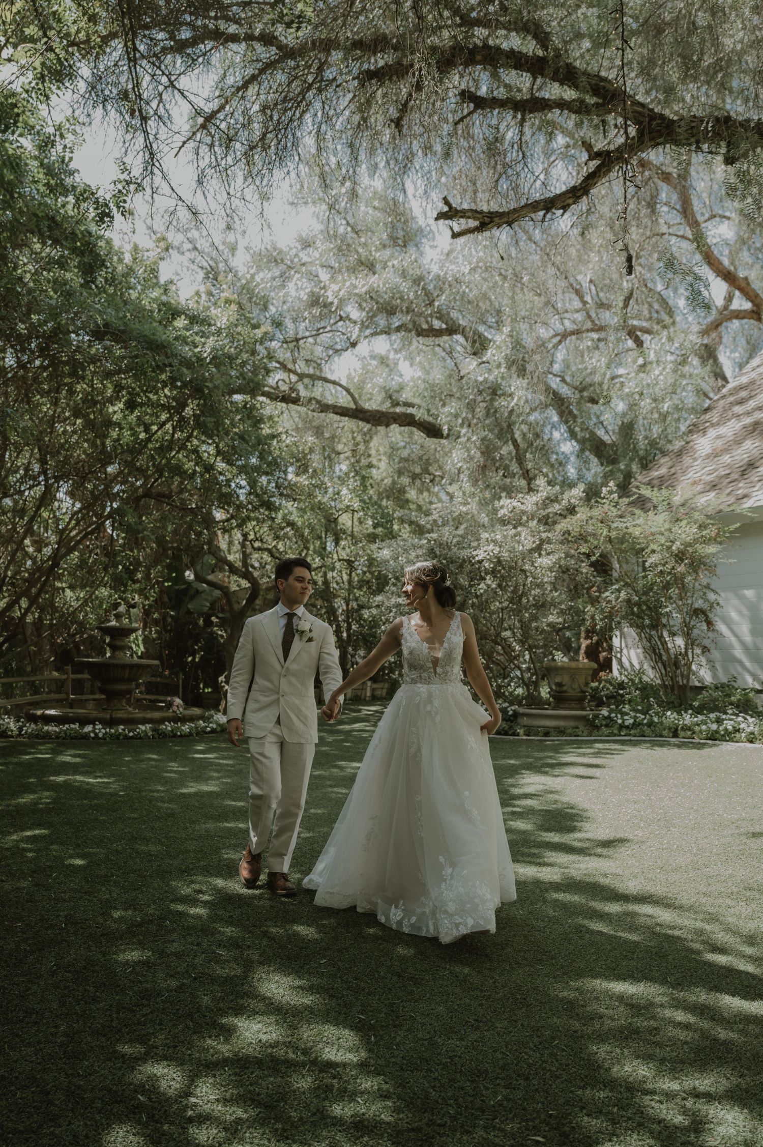 Bride and groom walk hand-in-hand through a lush garden, bride in floral lace gown, groom in beige suit.