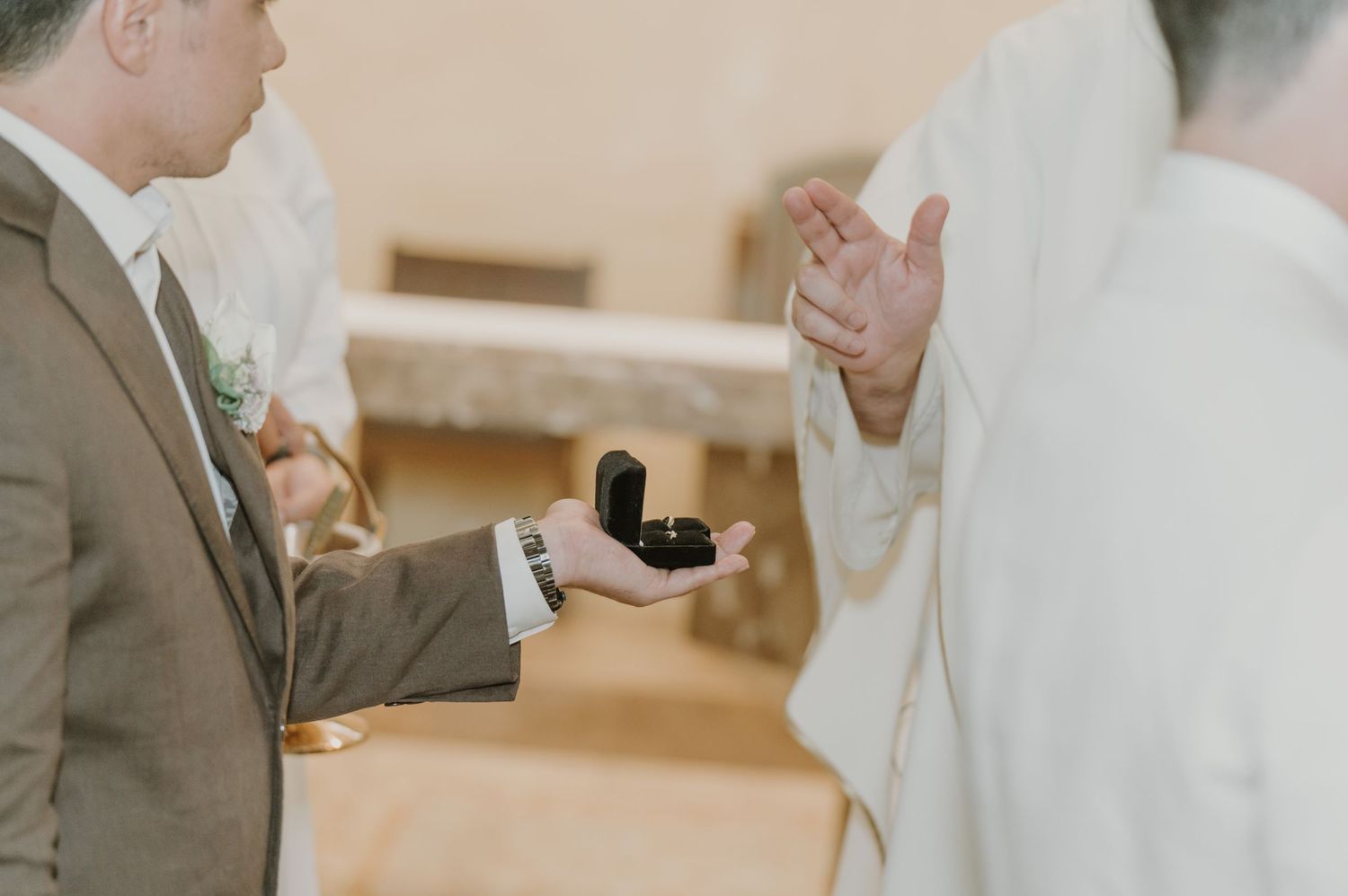 Groom holds open ring box as officiant blesses wedding rings during church ceremony.