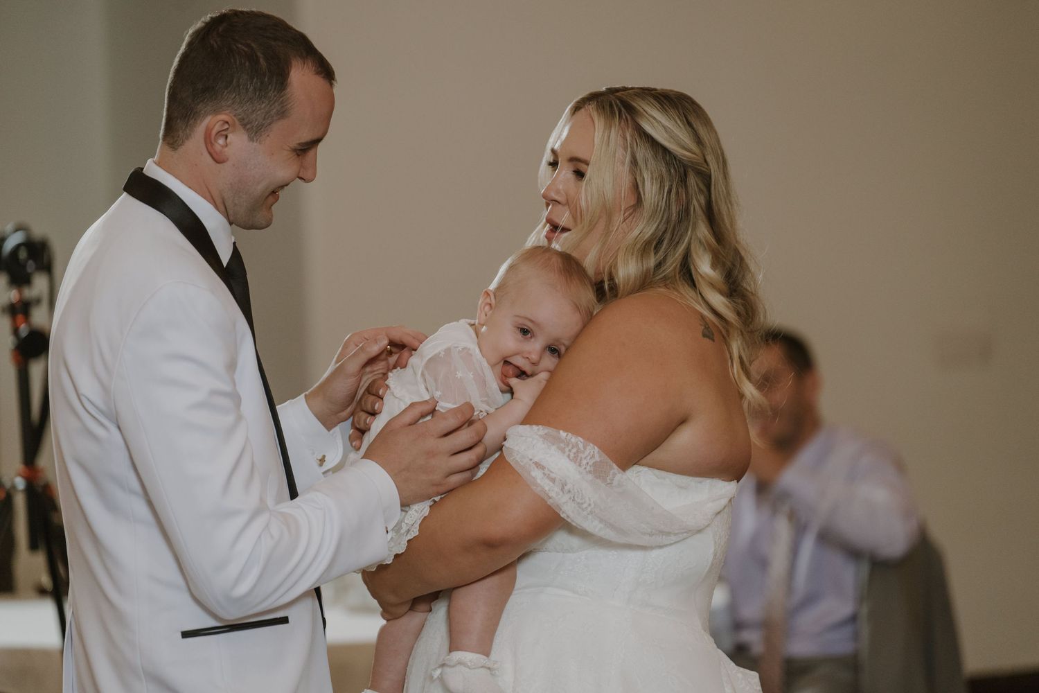 Bride and groom in white wedding attire smiling and holding a happy baby during their wedding ceremony.