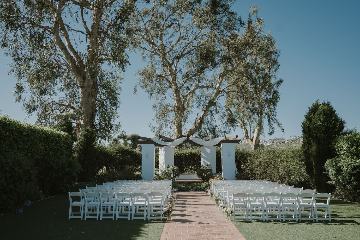 Outdoor wedding ceremony setup with white chairs, floral arch, and tall trees under a clear blue sky.