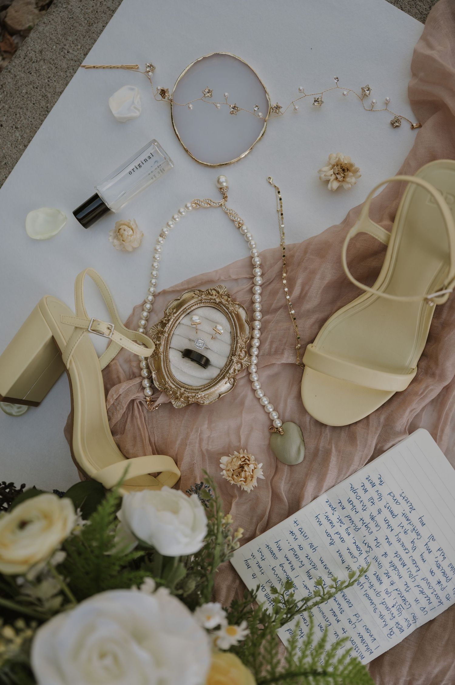 Wedding accessories including shoes, pearls, and handwritten notes laid out on blush fabric.