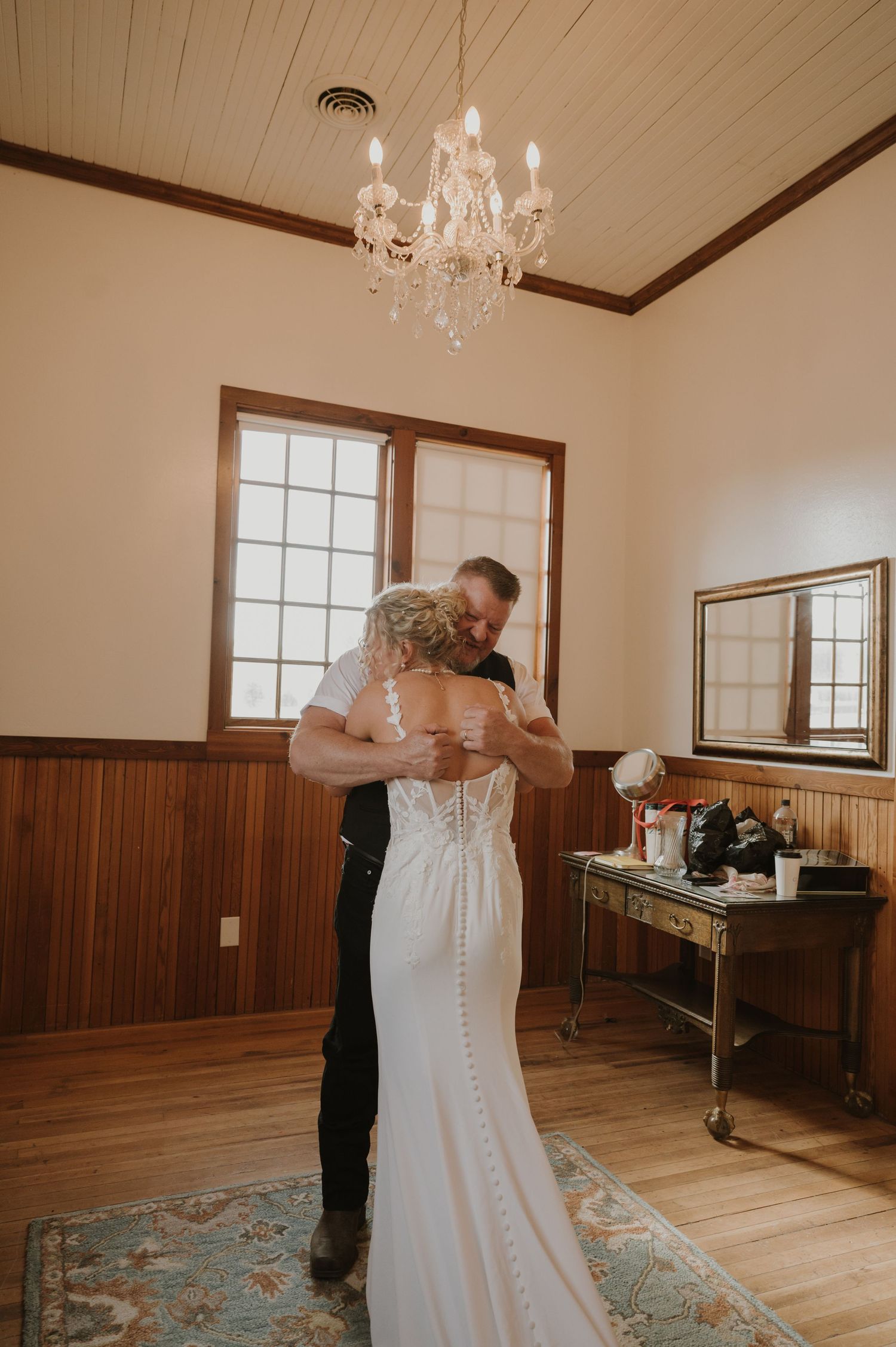 A wedding dress being buttoned up in a room with wood paneling and a crystal chandelier.