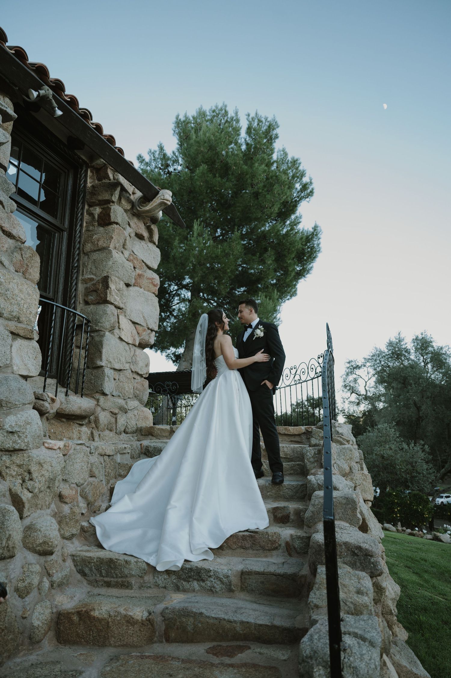 Couple in wedding attire pose on stone steps of rustic building at sunset.