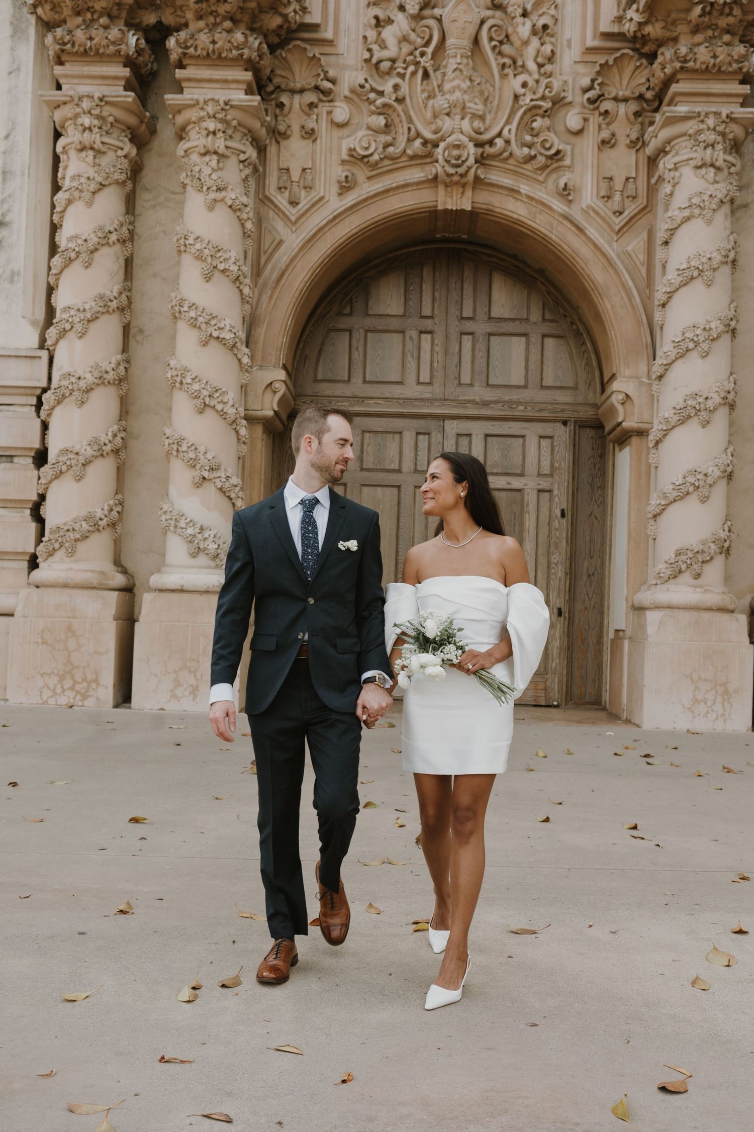Elegant wedding couple walks together near ornate stone columns and archways.